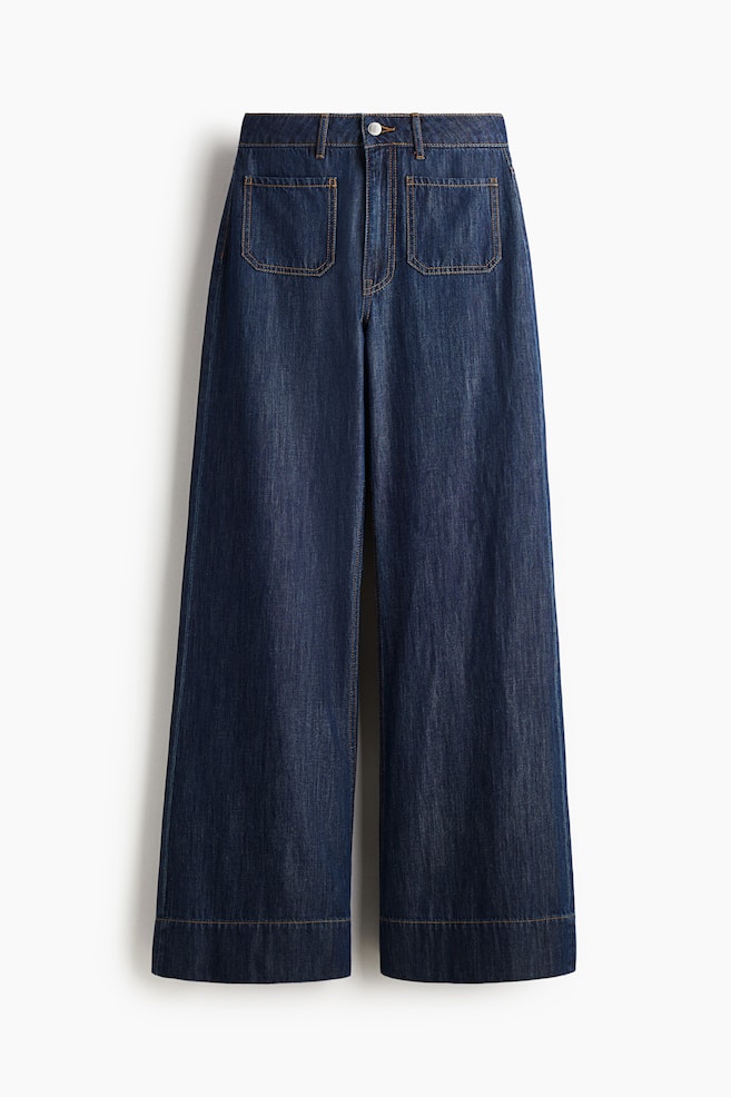 Vorderansicht von dunkelblauen, festen Denim-Jeans, die eine extra hohe Taille mit einem Reißverschluss und Knopfverschluss aufweisen. Die Jeans sind mit einem weiten Bein, einer regulären Passform von der Taille bis zur Hüfte und einem weiten Schnitt vom Oberschenkel bis zum Saum gestaltet, mit vorderen aufgesetzten Taschen und einer längeren Länge, die einen Stapel-Effekt über dem Fuß erzeugt. - 1