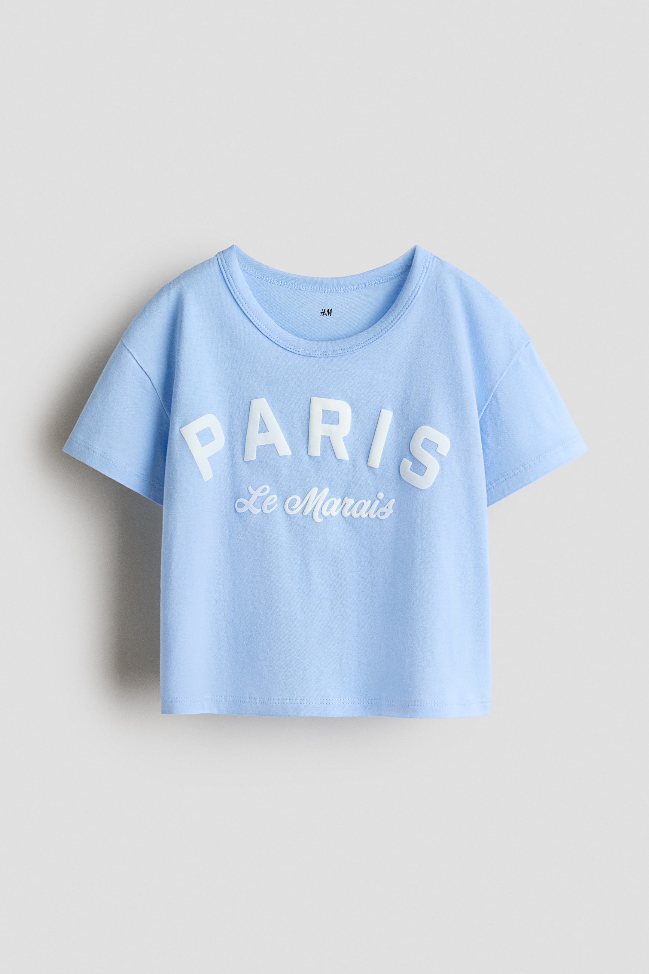 View larger image: A light blue short-sleeved t-shirt is presenting its FRONT, featuring a round neckline. It is displaying the word "PARIS" in large, raised white letters across the chest, with "Le Marais" in smaller, white script lettering positioned below.