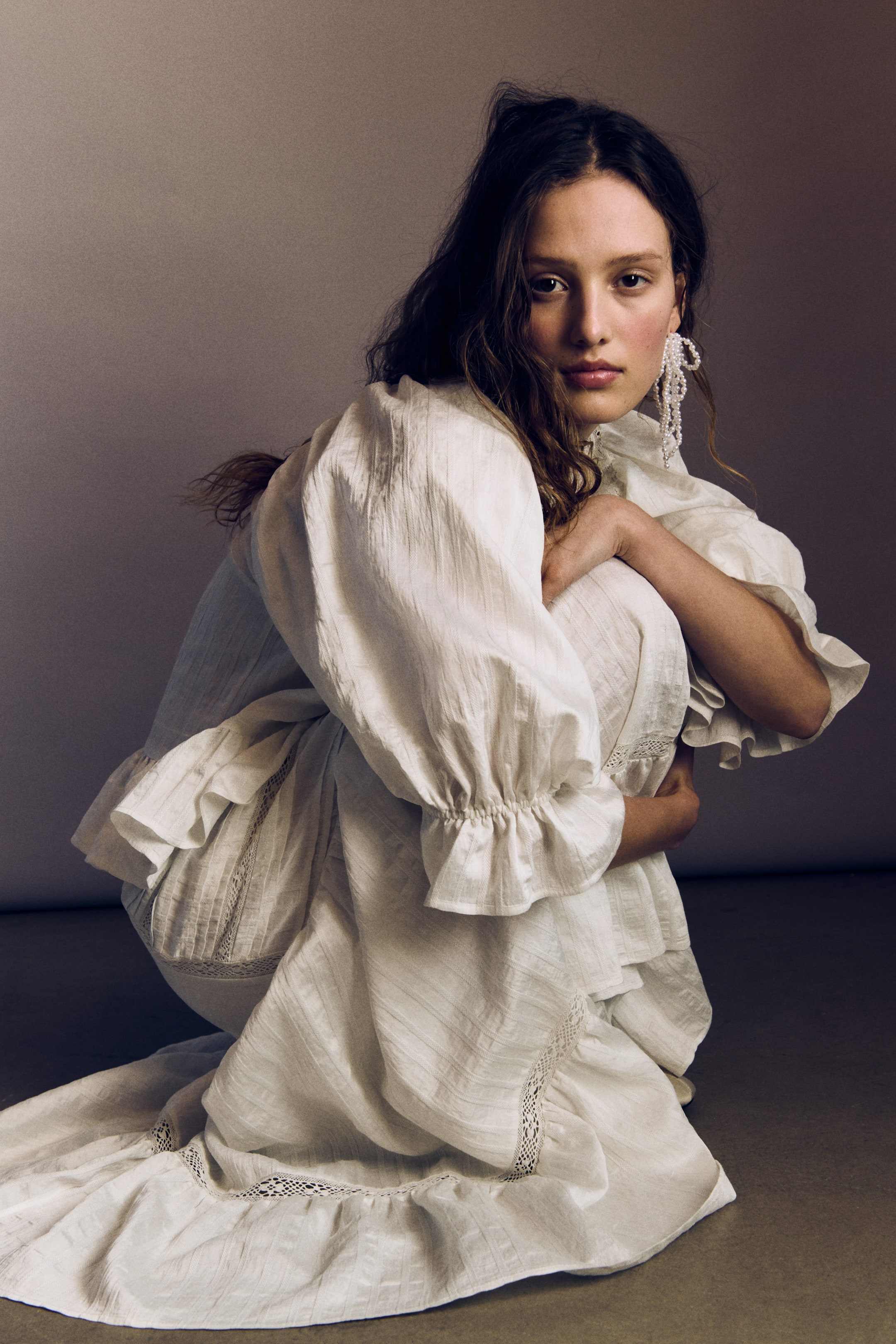 Agrandir l'image: Une robe à manches longues de couleur blanc cassé présente des manches bouffantes volumineuses avec des poignets à volants et un col montant à volants. La robe affiche une texture froissée avec de subtiles rayures verticales et des détails en dentelle sur sa jupe à étages avec un ourlet à volants, sa silhouette complète étant mise en valeur par la pose accroupie.