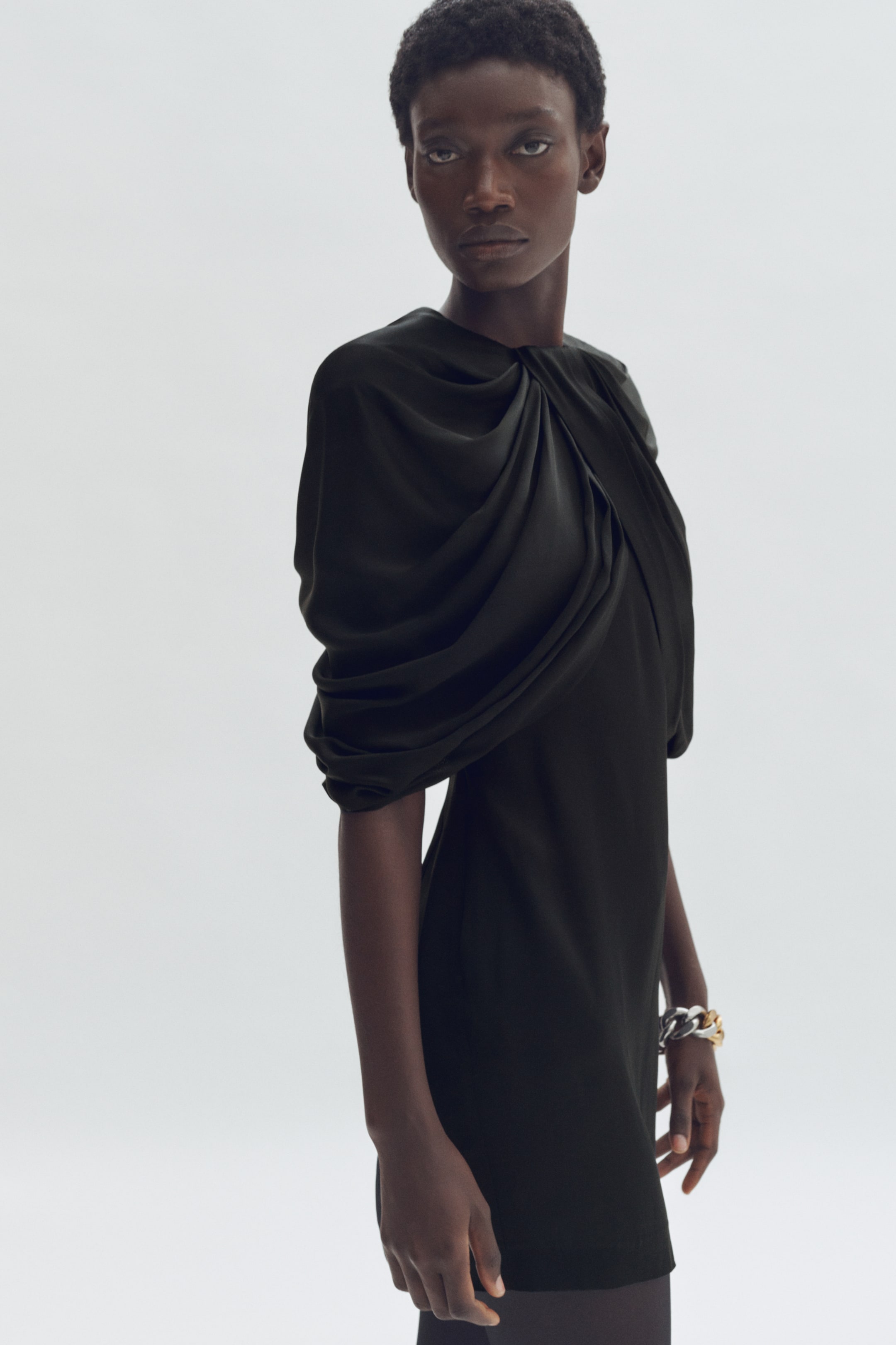 Voir une image plus grande: Une robe vert foncé présente un col drapé et torsadé sur le côté gauche, s'étendant en une manche courte volumineuse. La robe arbore une coupe droite et expose son profil avant et gauche, se terminant au-dessus du genou.