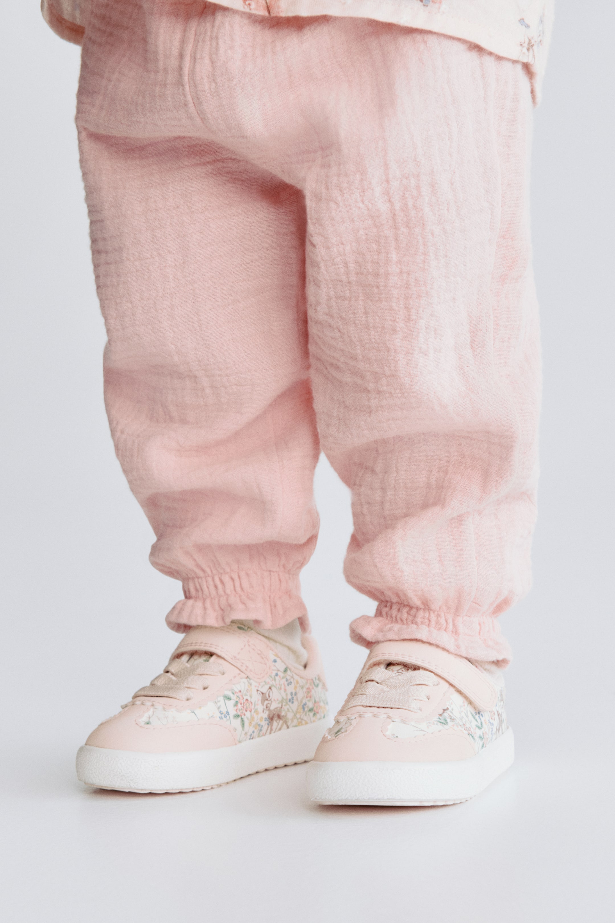 Visualizza immagine più grande: I pantaloni rosa stropicciati presentano polsini arricciati ed elasticizzati alle caviglie. Scarpe da ginnastica rosa pallido con motivi floreali e cinturini a strappo sono indossate, viste da un angolo leggermente frontale.