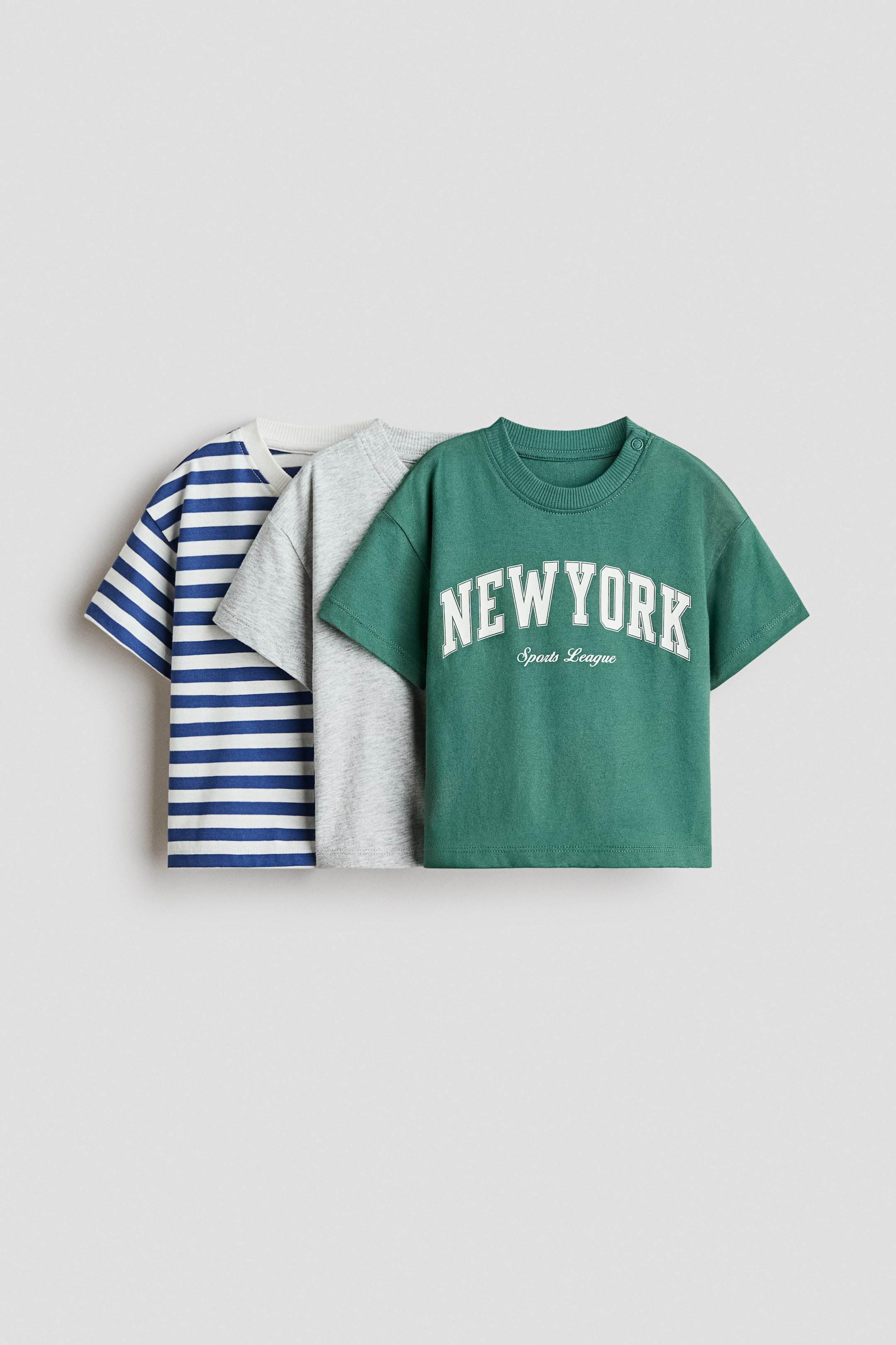 View larger image: A 3-pack of short-sleeved cotton T-shirts is overlapping and angled to the right, each featuring a ribbed crew neck. The first T-shirt is patterned with blue and white horizontal stripes, the second is heather grey, and the third is dark green, displaying "NEW YORK Sports League" text on its front and two snaps on the left shoulder.