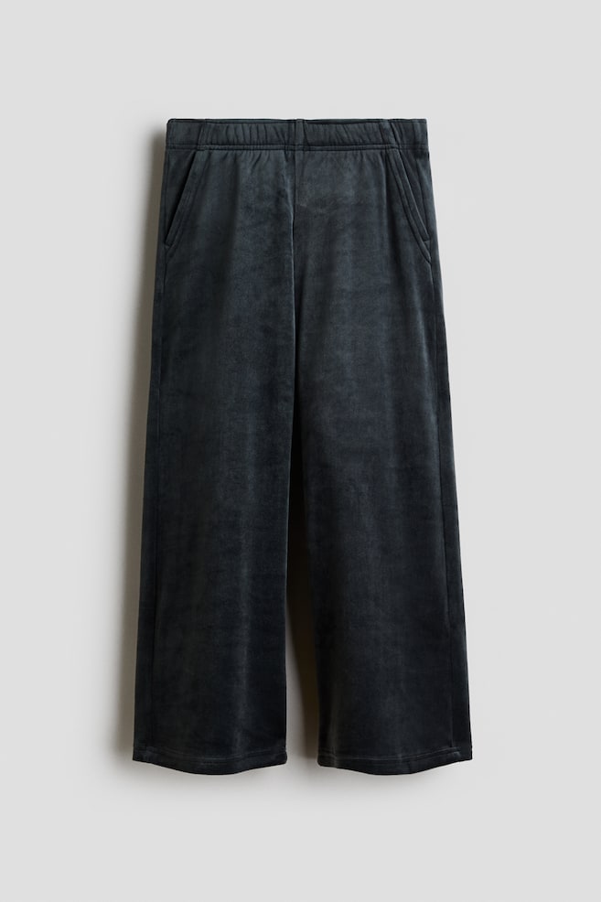 A front view of dark grey velour joggers displaying an elasticated waistband, diagonal side pockets, and straight legs. The pants are hanging flat to showcase their soft texture and relaxed structure. - 1