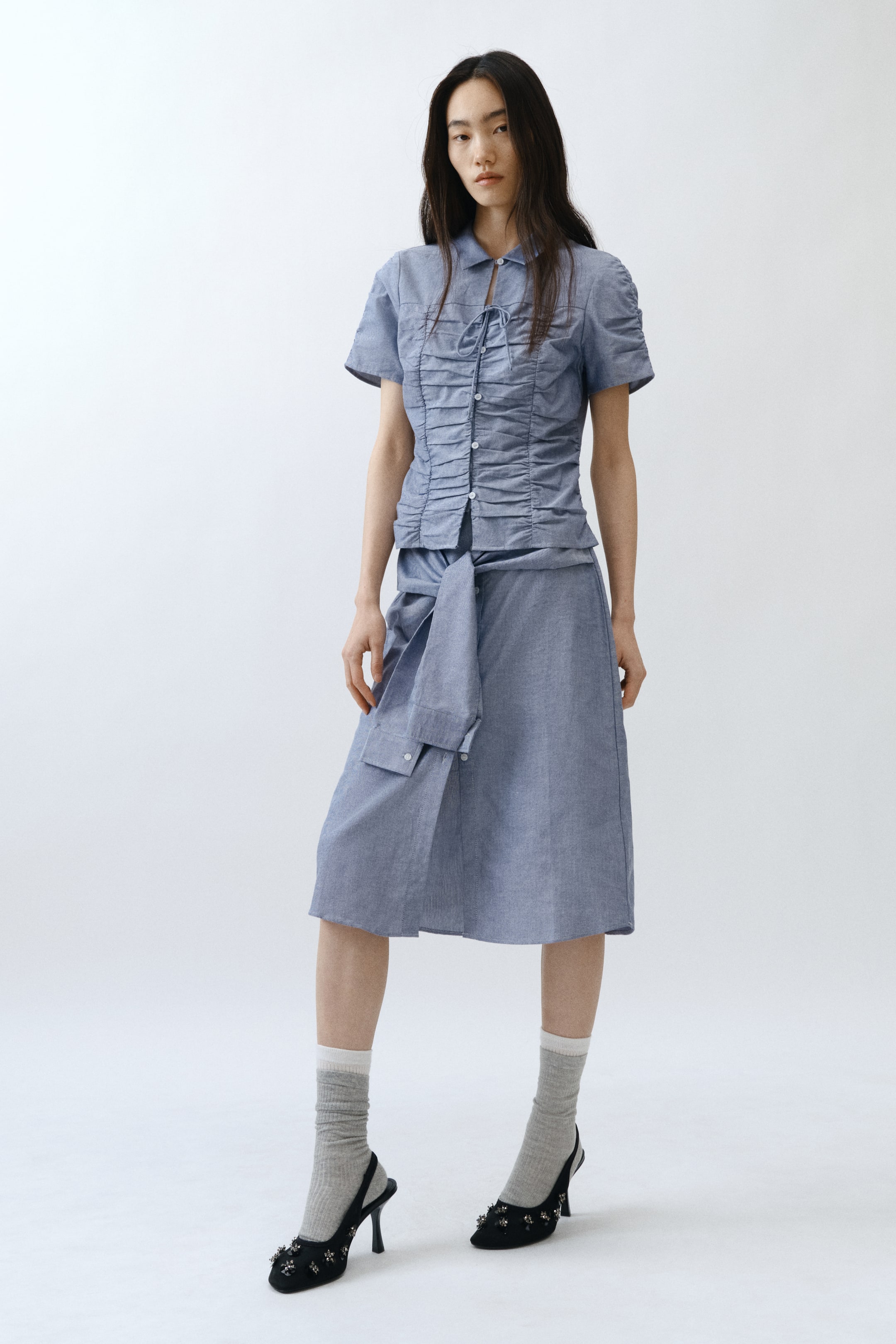 拡大画像を表示: ライトブルーのシャンブレー襟付き半袖トップスは、結び目のあるネックラインとギャザーが入ったフロントパネルが特徴です。それにマッチするミディスカートは、シャツの袖を結んだようなウエストタイディテールが施されています。このセットアップは、グレーのリブクルーソックスとブラックの装飾付きスリングバックヒールと共にスタイリングされています。