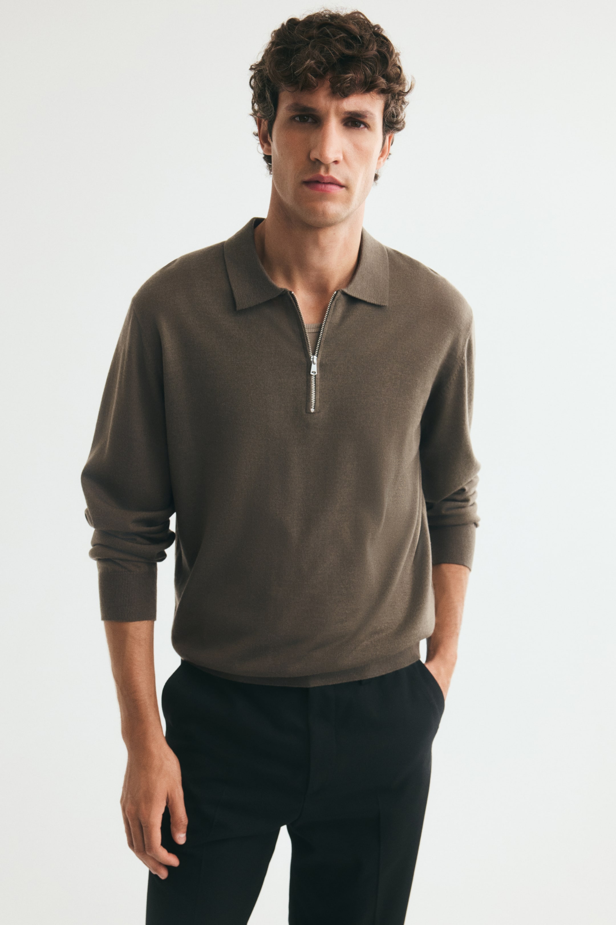 View larger image: A brown, fine-knit polo shirt featuring a classic collar, an open silver-tone half zip, and long sleeves with ribbed cuffs is being worn by the model. The model is pairing the regular-fit polo with black trousers, standing with one hand resting in a pocket.