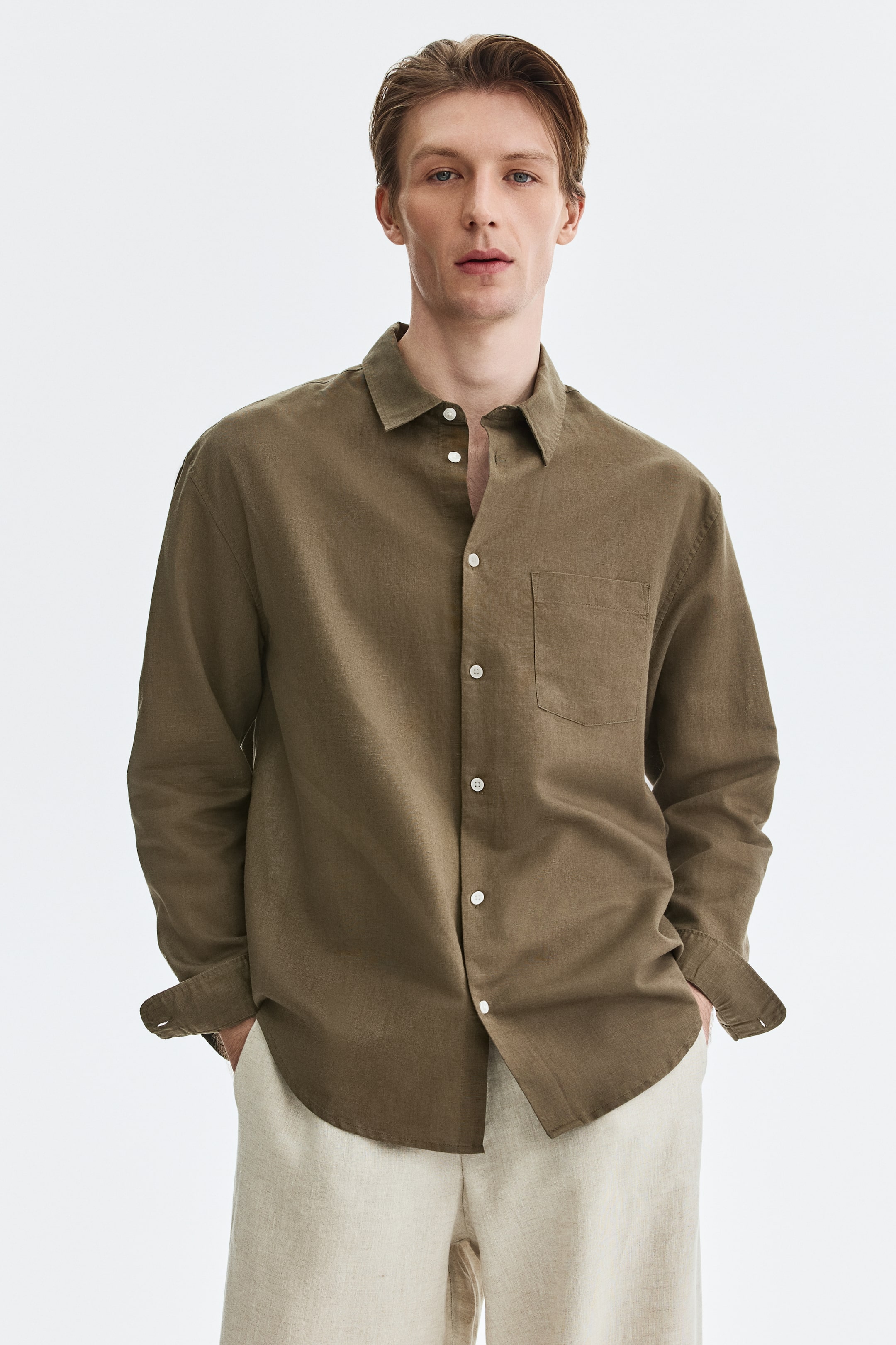 Ver imagen más grande: Una camisa de manga larga en color verde oliva está de frente, con un cuello abierto y botones blancos que recorren su tapeta frontal. Se aprecia un bolsillo de parche en el lado izquierdo del pecho, y los puños están doblados una vez, revelando un dobladillo curvado en los laterales inferiores.