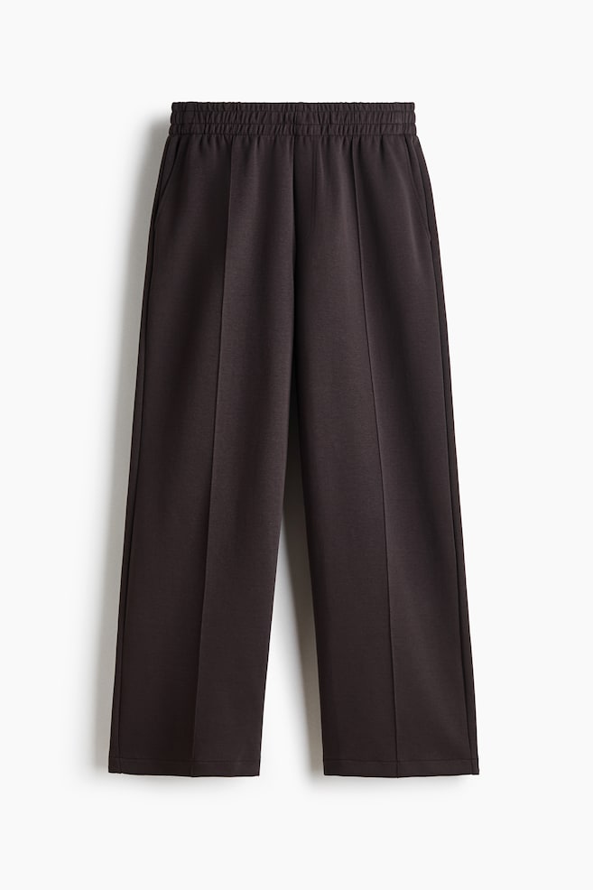 A pair of dark brown straight-leg trousers is hanging front-facing, featuring a gathered elasticated waistband and crisp pintuck creases running down the centre of each leg. The trousers showcase subtle side-seam pockets and a relaxed, structured drape. - 1