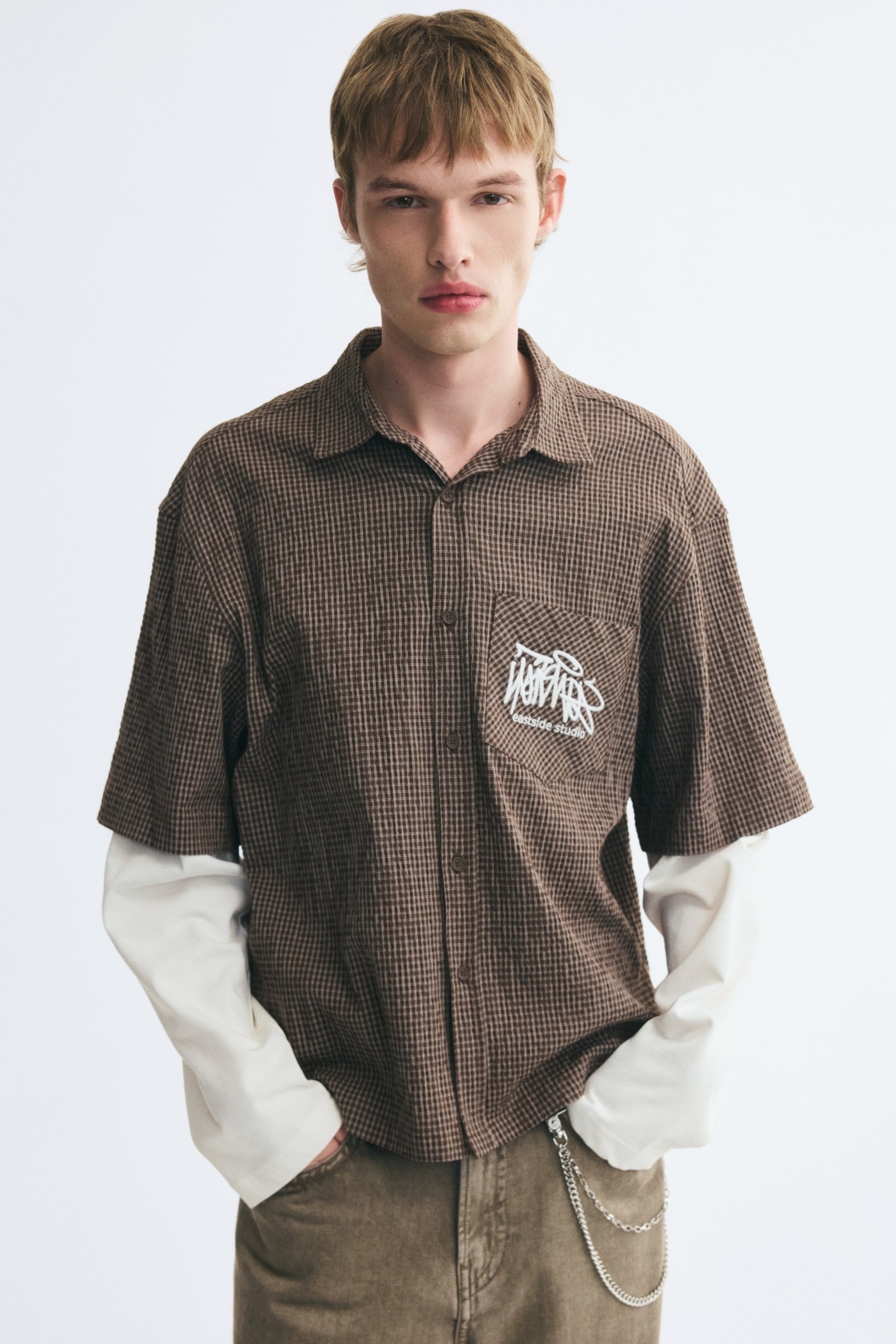 View larger image: A brown gingham-patterned short-sleeve shirt featuring a white embroidered graphic on the chest pocket is being worn by the model over integrated white long sleeves. The relaxed-fit collared shirt is styled with brown trousers.