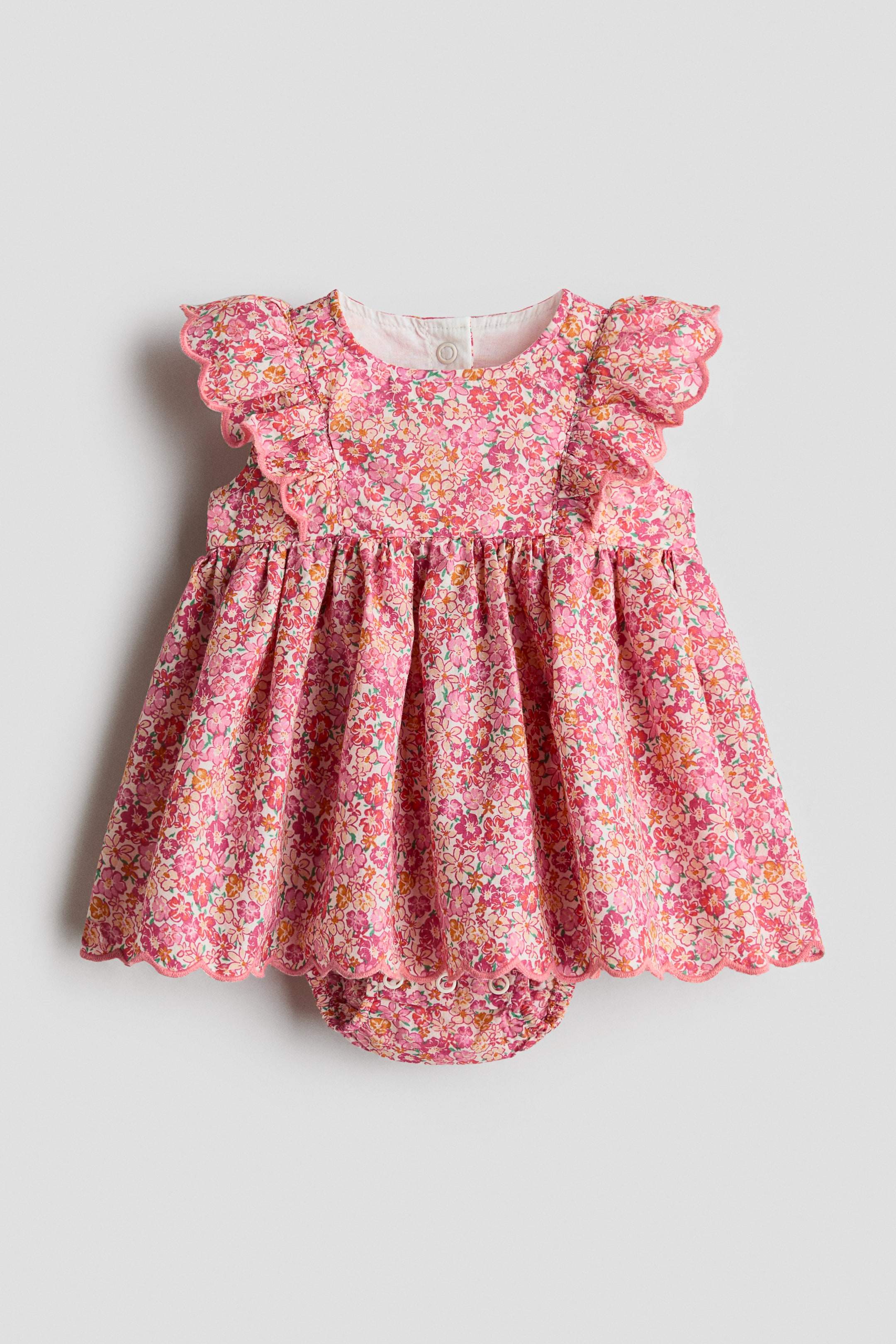 View larger image: The product, shown FRONT, is a sleeveless dress adorned with a pink floral design and an attached bodysuit. It showcases a round neckline, pink-edged scalloped frill trims that extend over the shoulders, a gathered seam that gives rise to a voluminous skirt, and an integrated bodysuit featuring press-studs at the bottom.