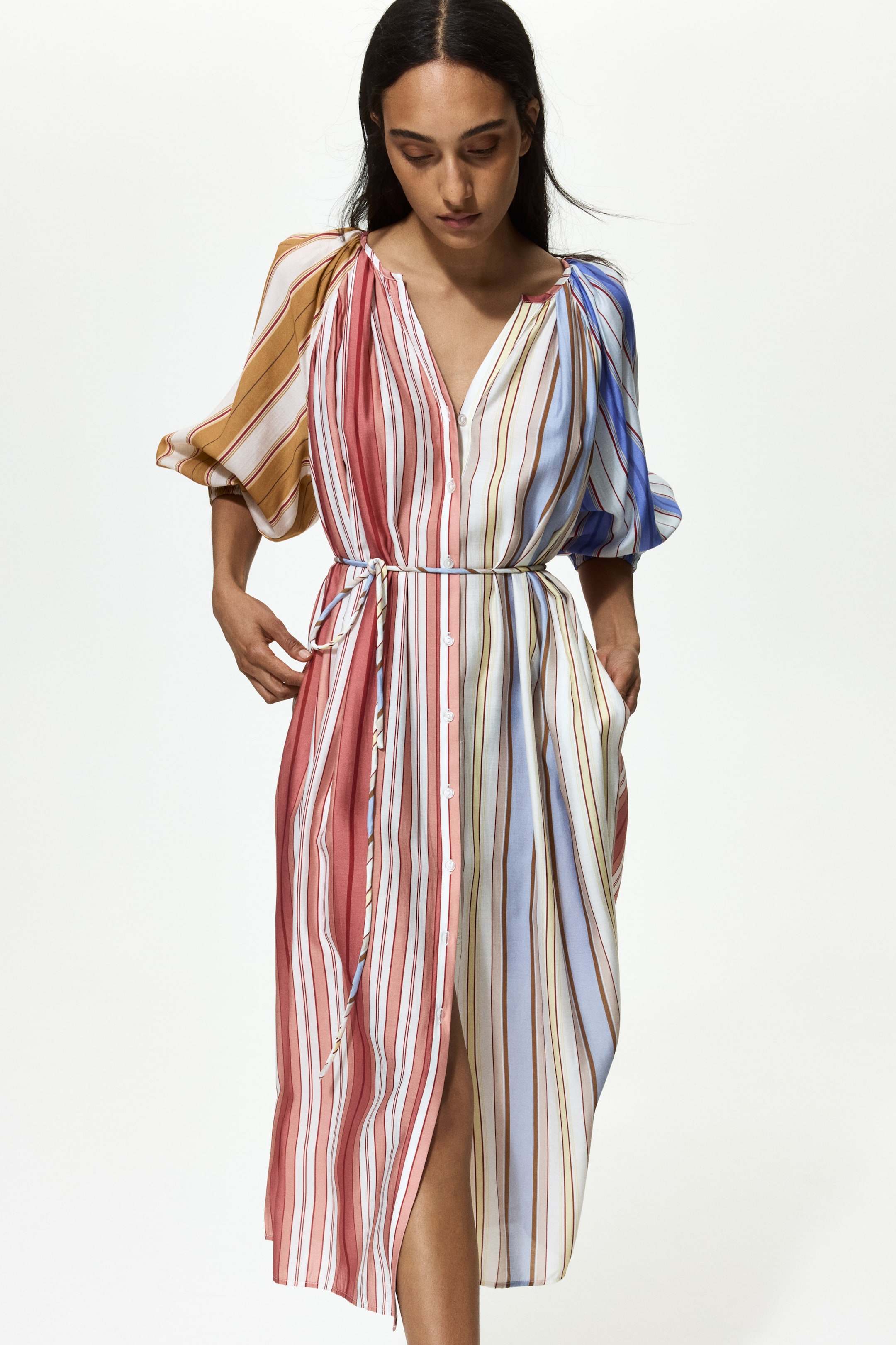 View larger image: A multi-colour striped midi dress is facing slightly right, featuring a V-neckline, puffed elbow-length sleeves, and a drawstring waist. It is showcasing a full-length front button placket, side pockets, and a high front slit, with varied vertical stripes in shades of red, blue, brown, yellow, and white.