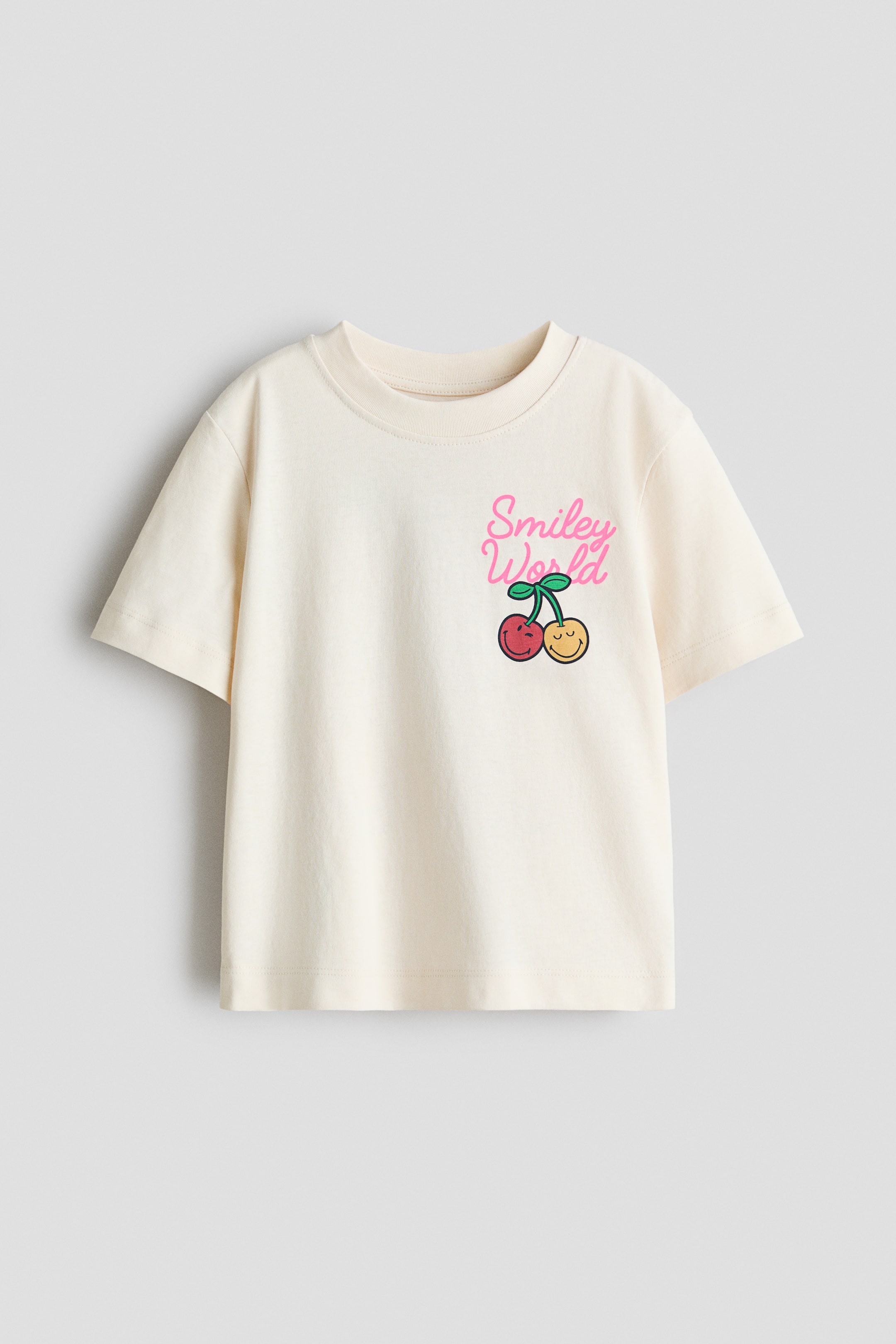 View larger image: A cream, loose-fit T-shirt is presenting its front, featuring a round, rib-trimmed neckline, short sleeves, and a straight-cut hem. A colourful graphic print is appearing on the left chest, displaying "Smiley World" in pink script above two cherries, one red with a smiling face and one yellow with a winking face.