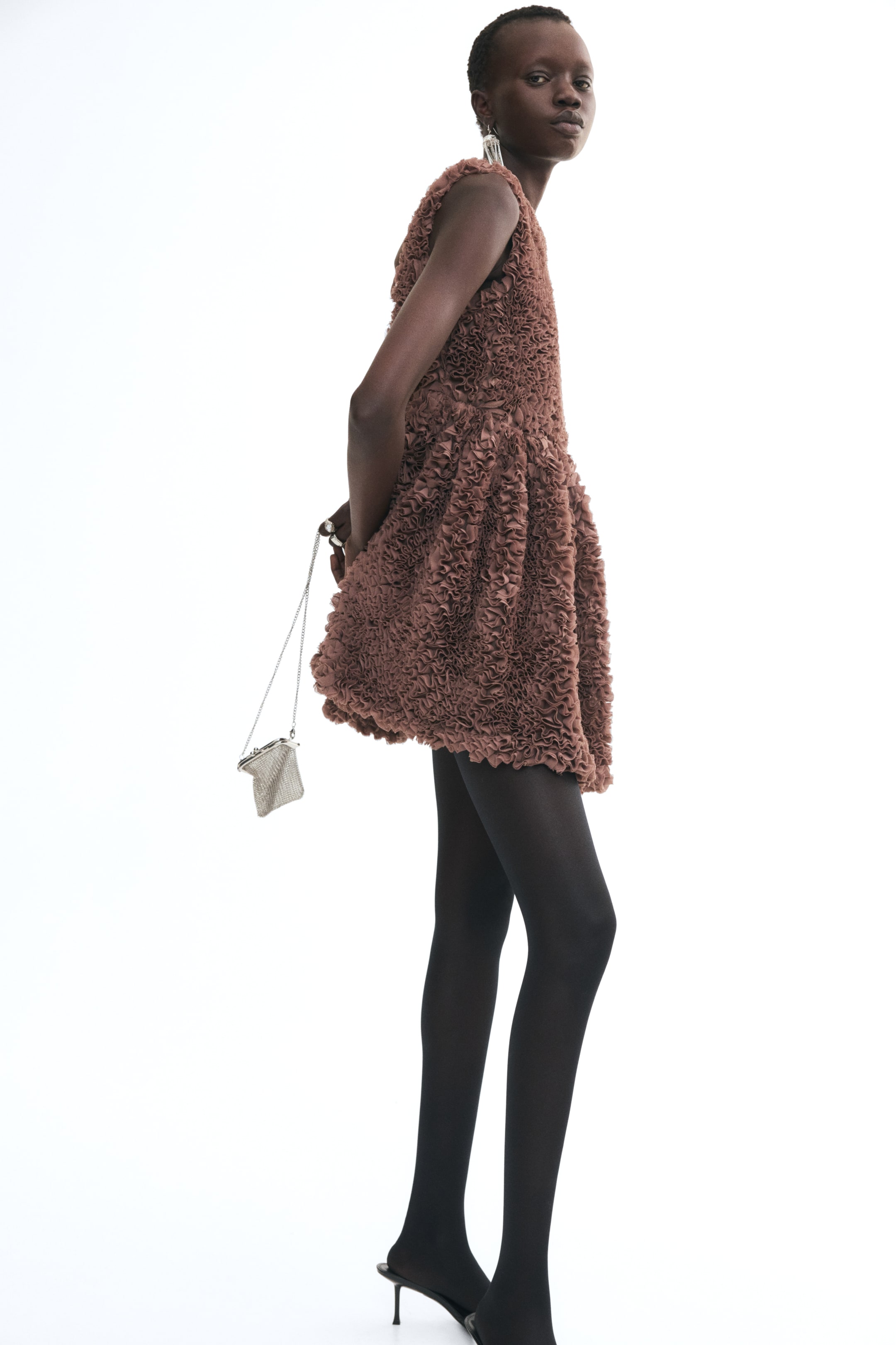Grösseres Bild ansehen: Ein strukturiertes, braunes ärmelloses Minikleid mit dichtem Rüschen-Detail wird vom Model getragen. Das Kleid wird mit schwarzen Strumpfhosen gestylt, und eine kleine silberne Kettenhandtasche wird hinter dem Rücken des Models gehalten.