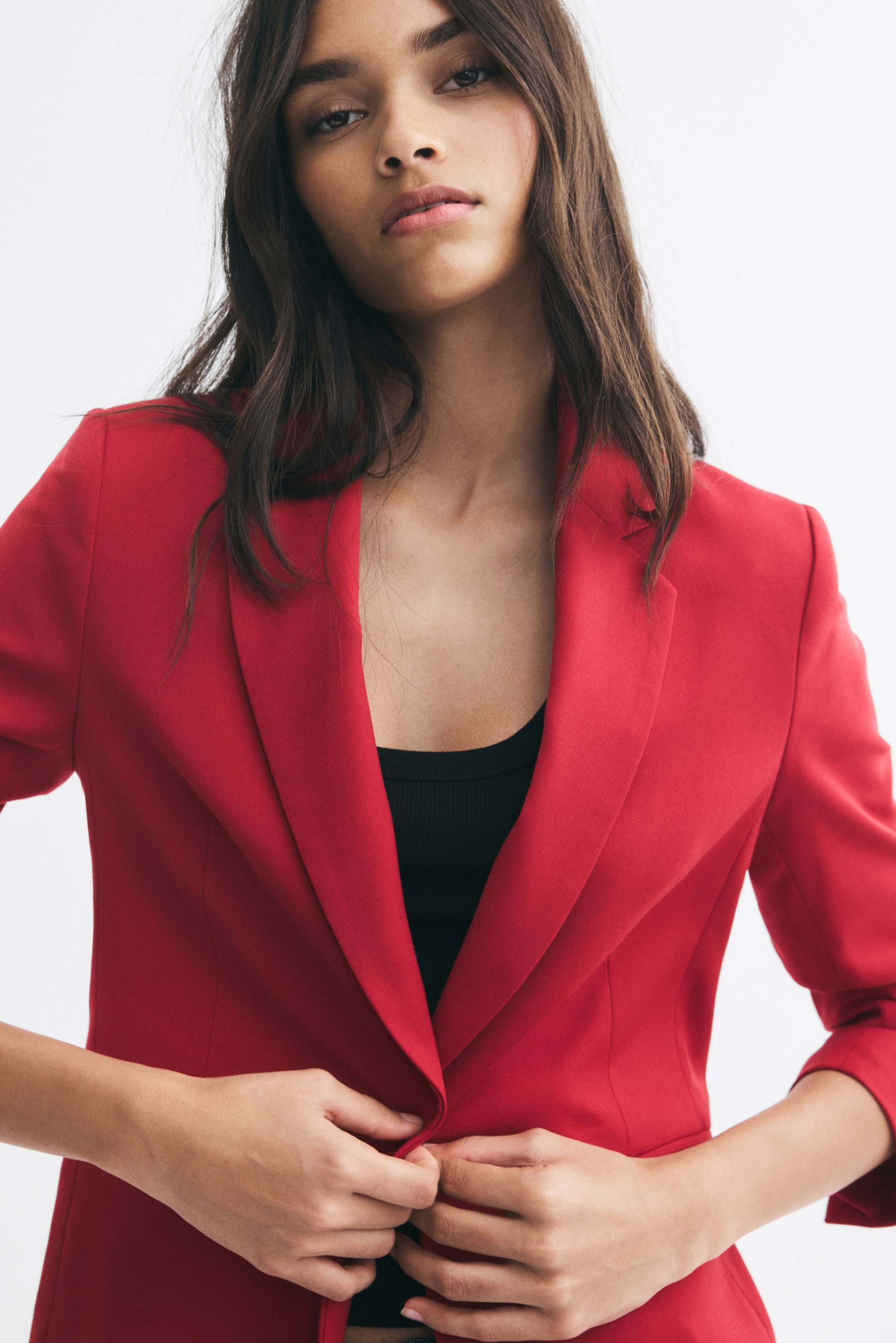View larger image: The model is wearing a red, single-breasted blazer with notch lapels over a black top. She is holding the front opening of the blazer together with both hands near the waist.