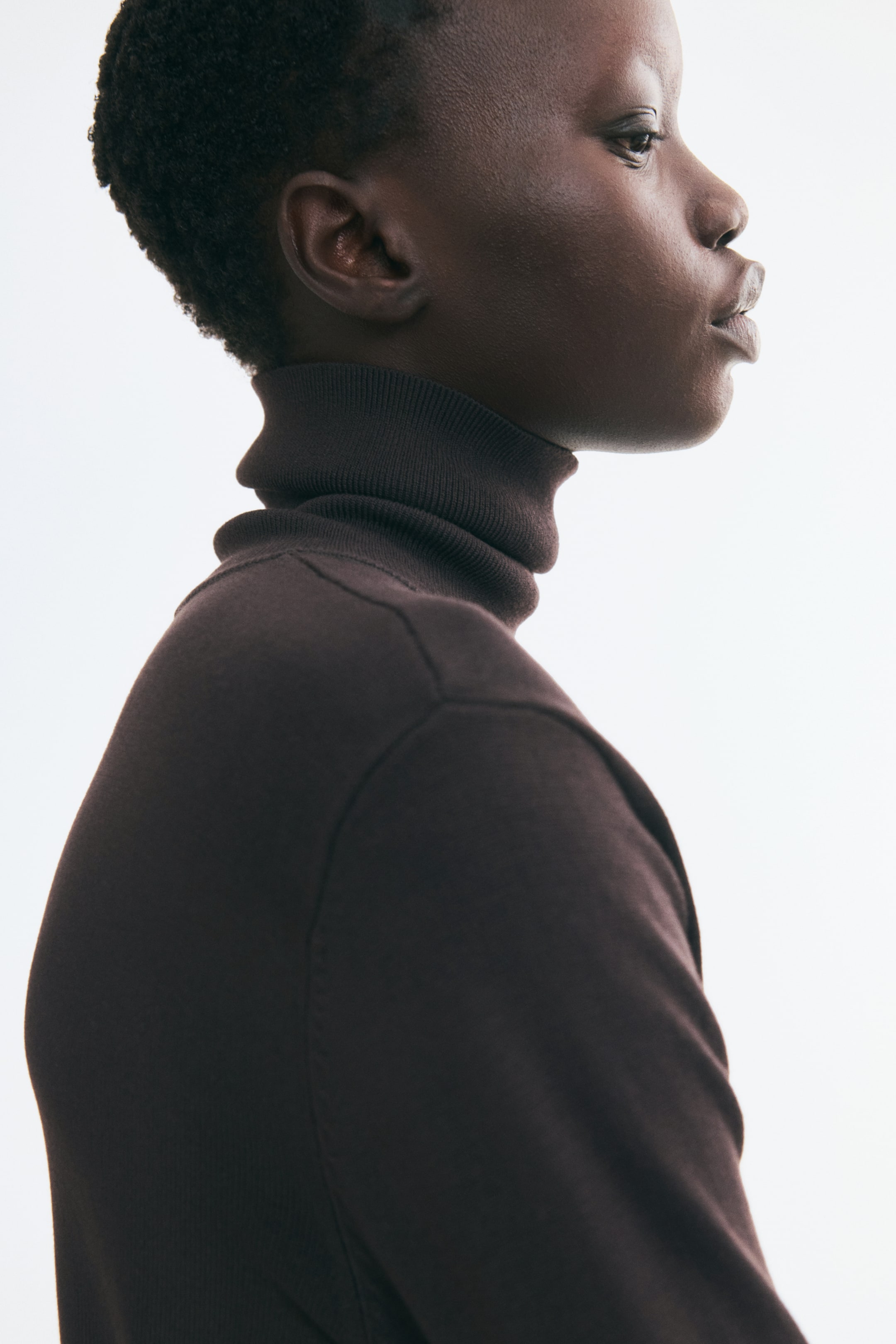 Grösseres Bild ansehen: Ein dunkelbrauner Feinstrickpullover mit umgeschlagenem, geripptem Rollkragen wird vom Model in einer seitlichen Profilansicht getragen. Die Nahaufnahme betont die weiche Textur des Stoffes und die Schulternaht des Kleidungsstücks.