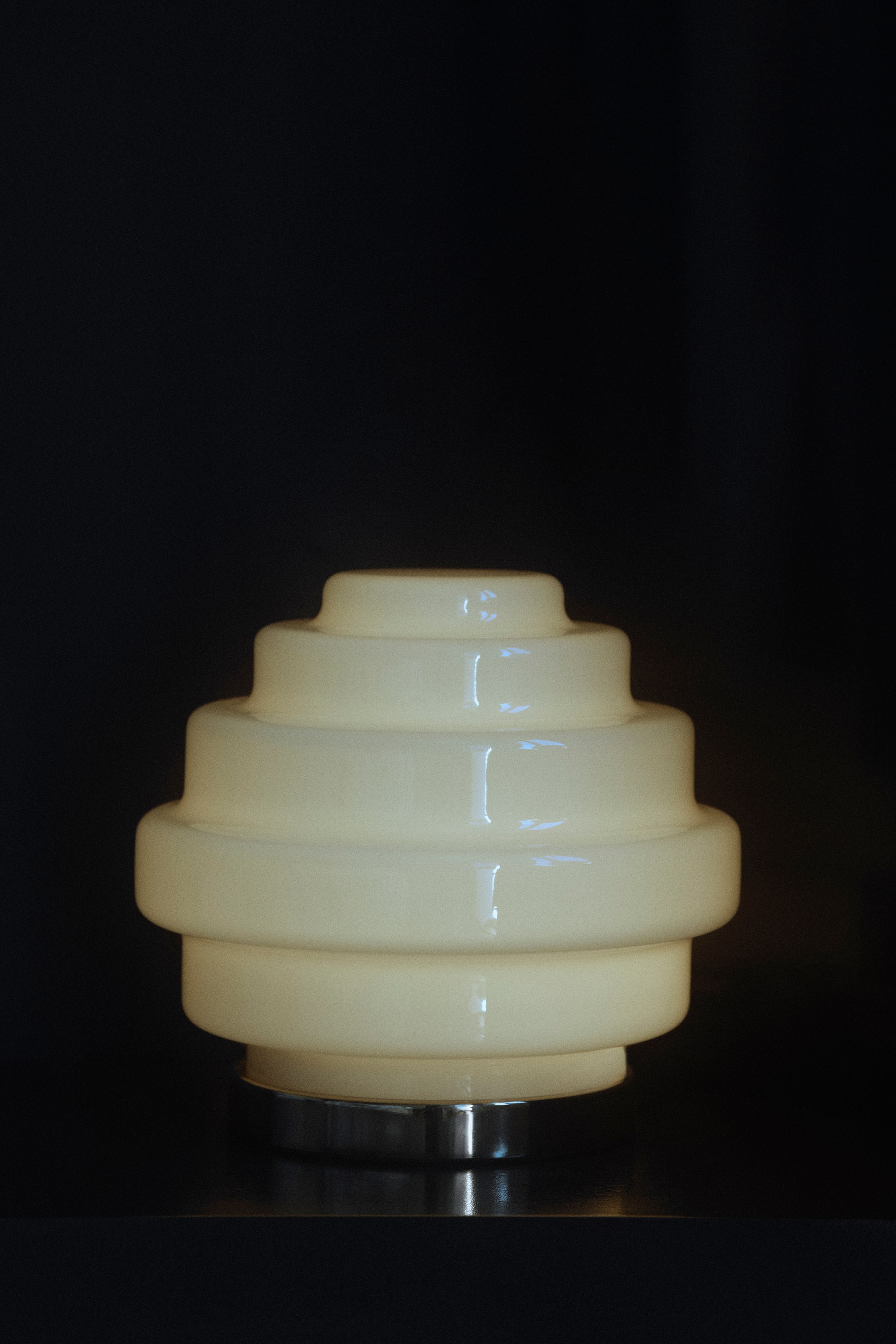 Grotere afbeelding bekijken: Een kleine tafellamp staat naar voren gericht en toont een glanzende, crèmekleurige glazen lampenkap met een gelaagd, getrapt geometrisch ontwerp. De lamp staat op een ronde, gepolijste metalen voet in zilverkleur en verspreidt van binnenuit een zachte, warme gloed.