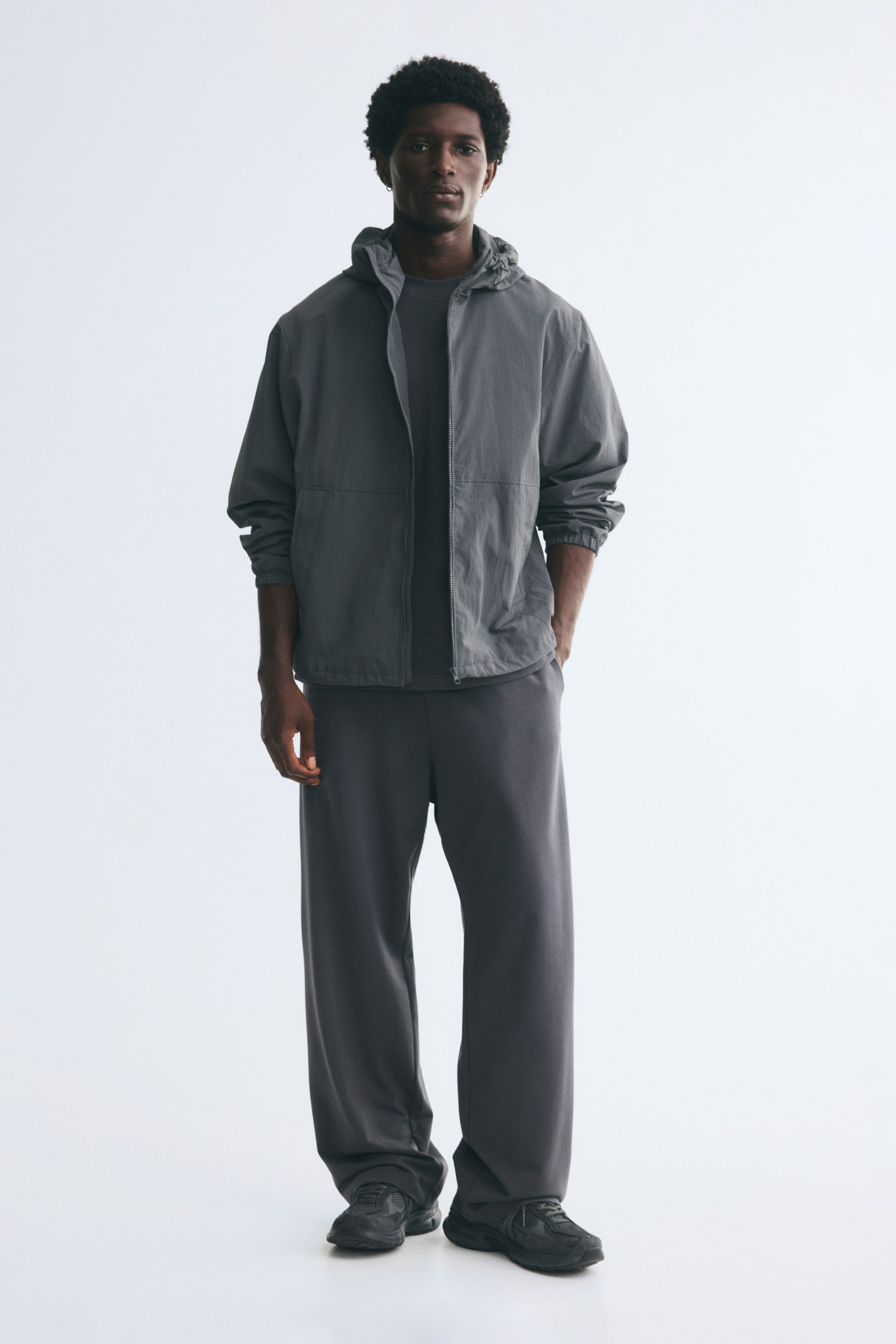 Voir une image plus grande: Le mannequin porte un pantalon de jogging ample en tissu molletonné gris foncé, debout avec une main posée dans une poche latérale. Le pantalon tombe souplement sur des sneakers foncées et est agencé avec un t-shirt assorti et une veste à capuchon grise à fermeture éclair.
