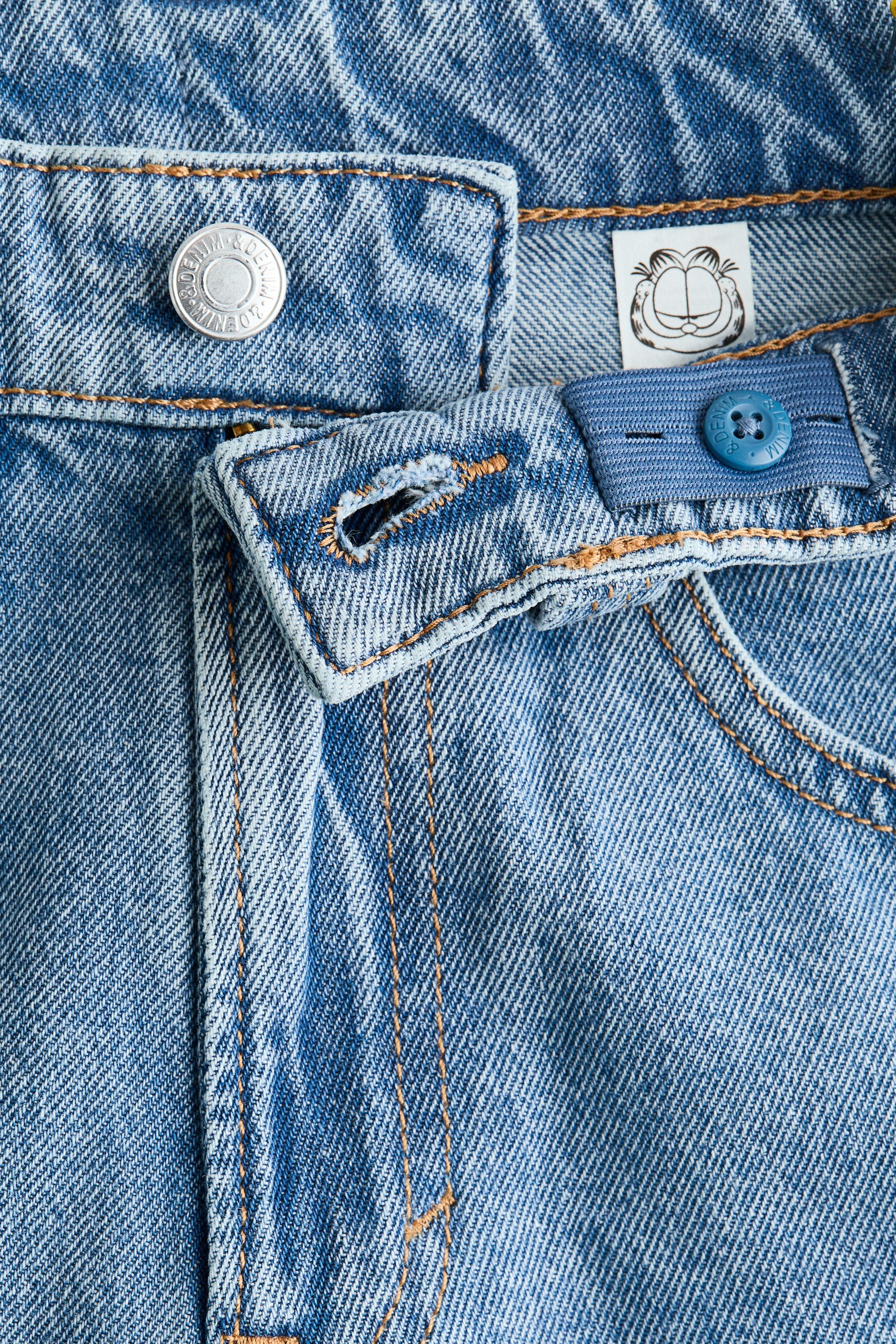 Agrandir l'image: FRONT. Un jean en denim bleu clair affiche les détails de sa taille et de sa fermeture. Un bouton en métal argenté estampillé "H&M DENIM" sécurise la ceinture, tandis qu'un onglet à bouton bleu réglable et élastique, également équipé d'un bouton "H&M DENIM", est visible à l'intérieur de la ceinture, à côté d'une étiquette en tissu blanc illustrant Garfield.