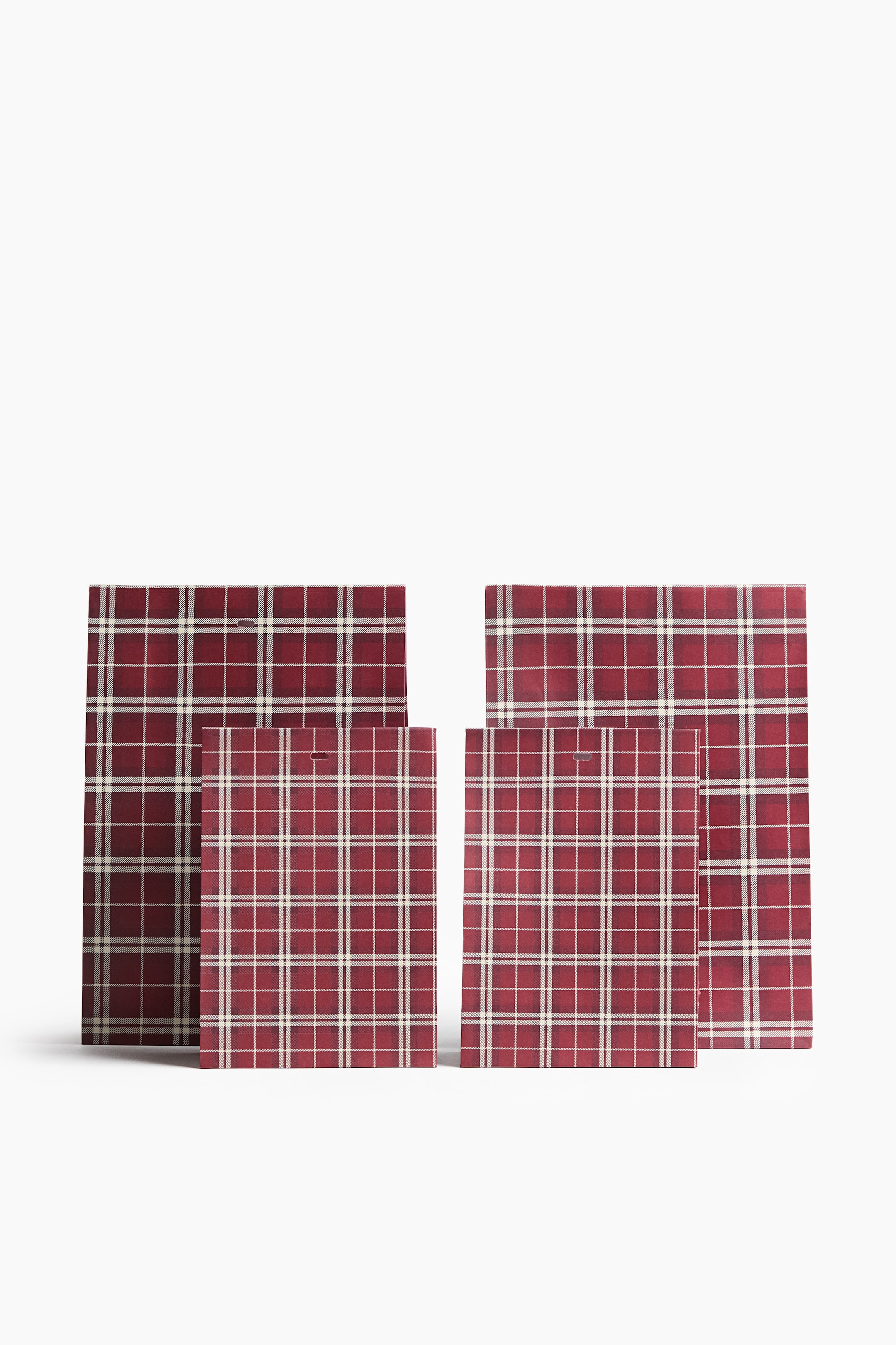 View larger image: A set of four paper gift bags, comprising two larger and two smaller sizes, stands upright facing forwards. Each bag is decorated with a red, white and beige plaid pattern and features a small pre-punched horizontal slot near the top centre.