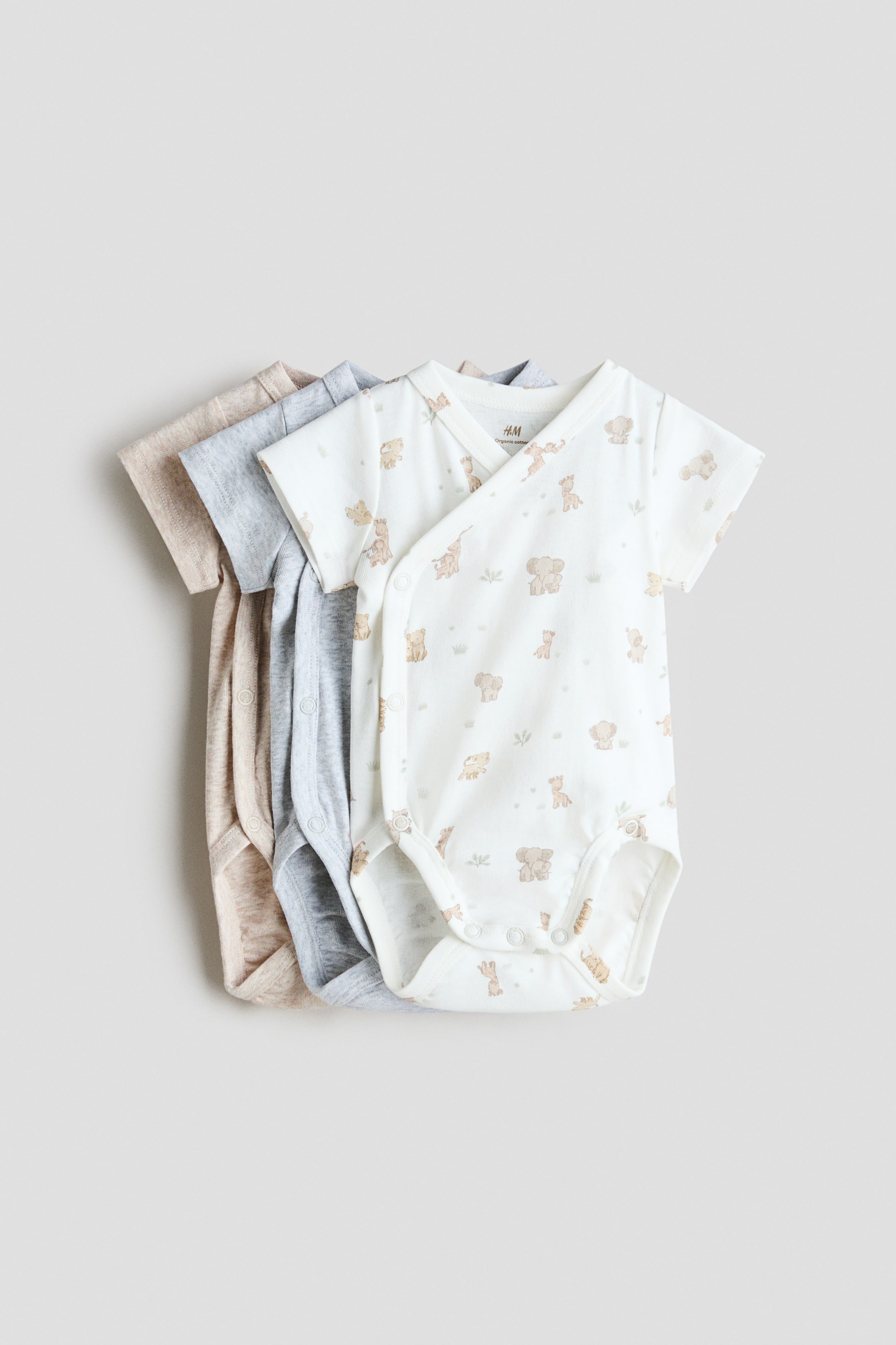 拡大画像を表示: 柔らかなコットンジャージー製のショートスリーブのラップオーバー式ボディスーツが三着、重ねて置かれています。手前のボディスーツはベビー動物柄の白地で、片側と股下にスナップ留めが付いているのが見えます。その下には無地のライトグレーとライトベージュのボディスーツが重ねられています。