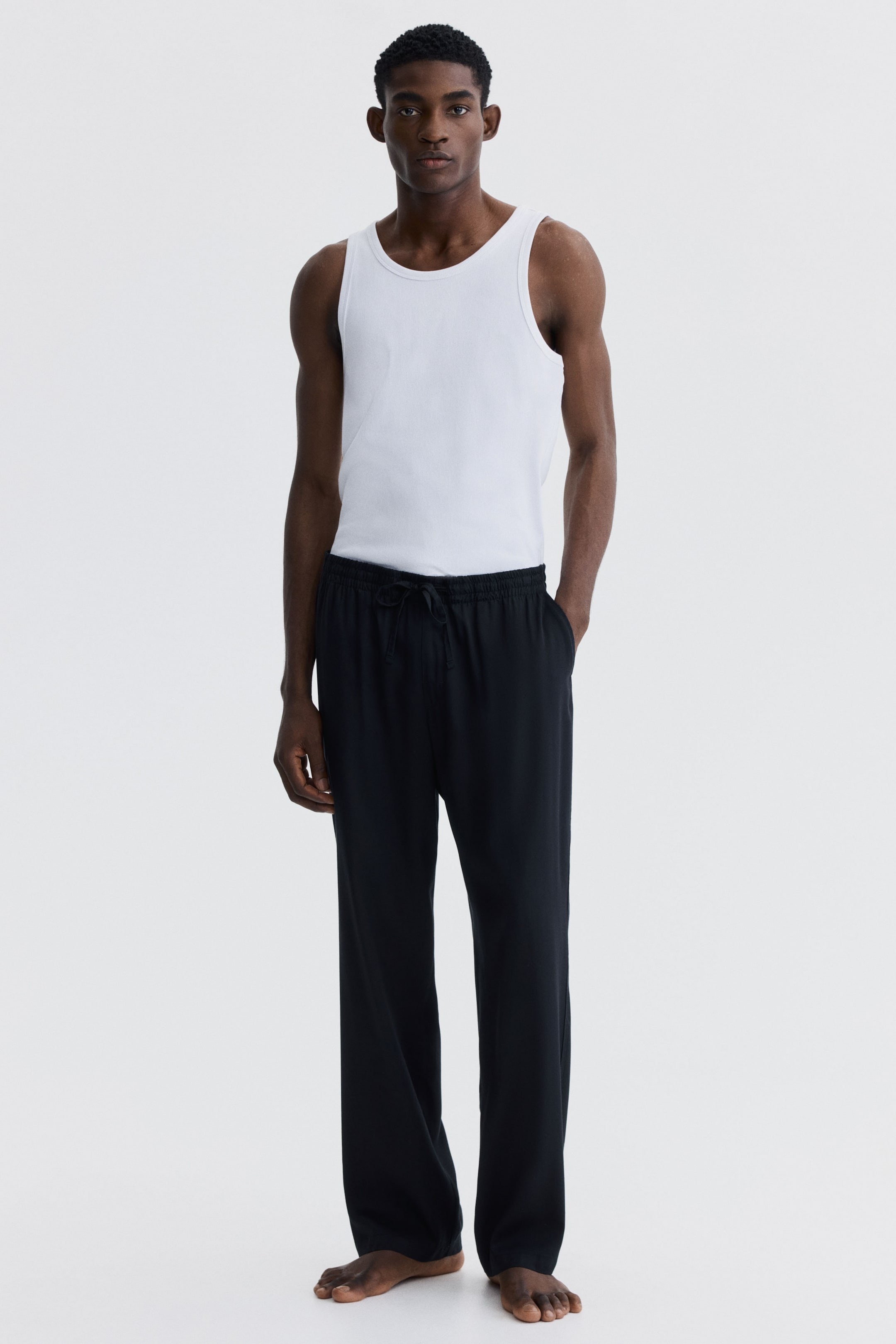 View larger image: Black trousers offer a relaxed, straight-leg fit with an elasticated drawstring waistband tied at the front, and side pockets, falling loosely to the ankles.