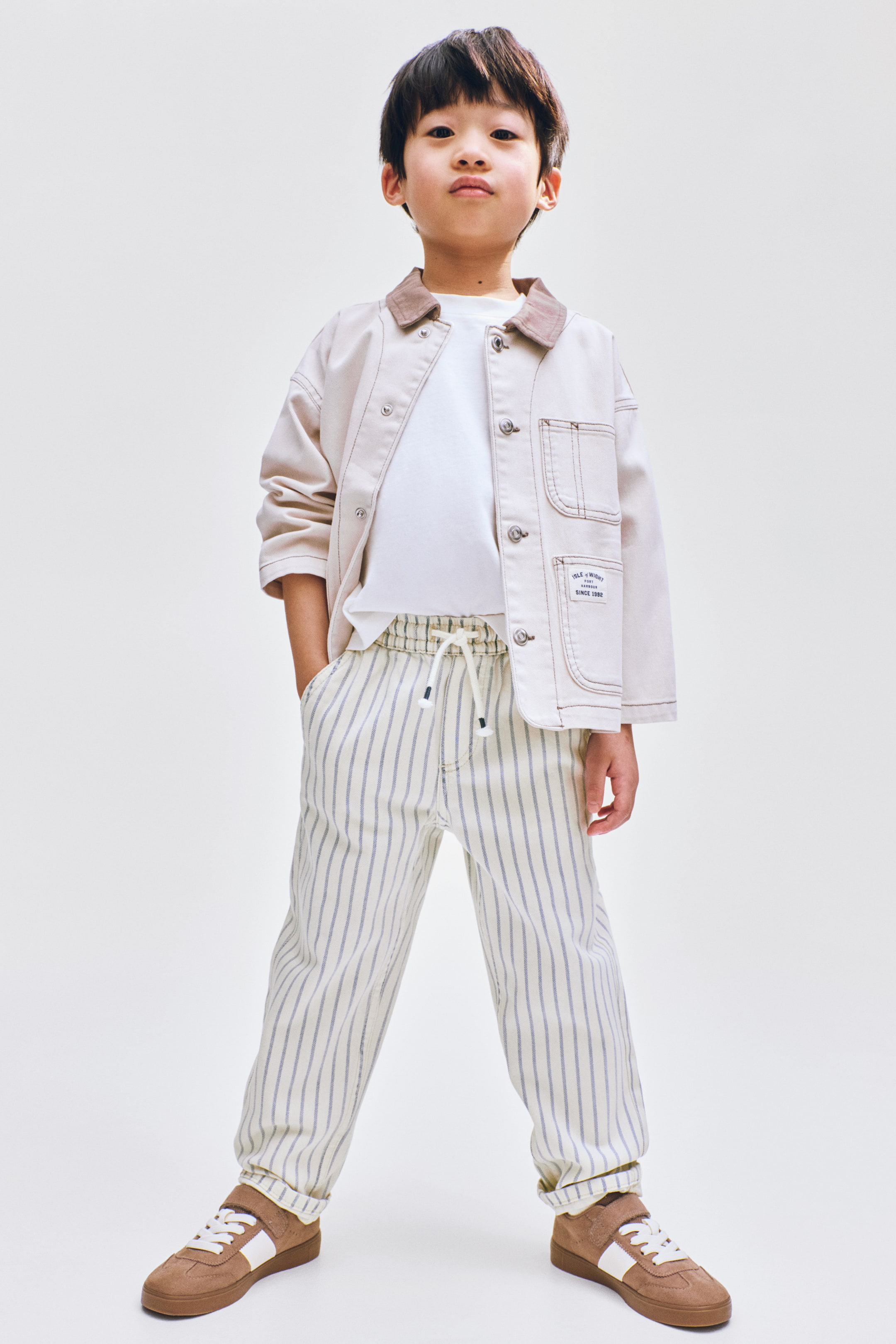 הצגת תמונה גדולה יותר: ז'קט עבודה בצבע בז' בהיר עם צווארון חום, כפתורי לחיצה ותפירה בולטת נלבש מעל חולצת טריקו לבנה. מכנסי קרם עם פסים אנכיים דקים, מותן עם גומי ושרוך, ושולי מכנסיים מקופלים נלבשים עם נעלי סניקרס חומות עם פסים לבנים בצדדים.