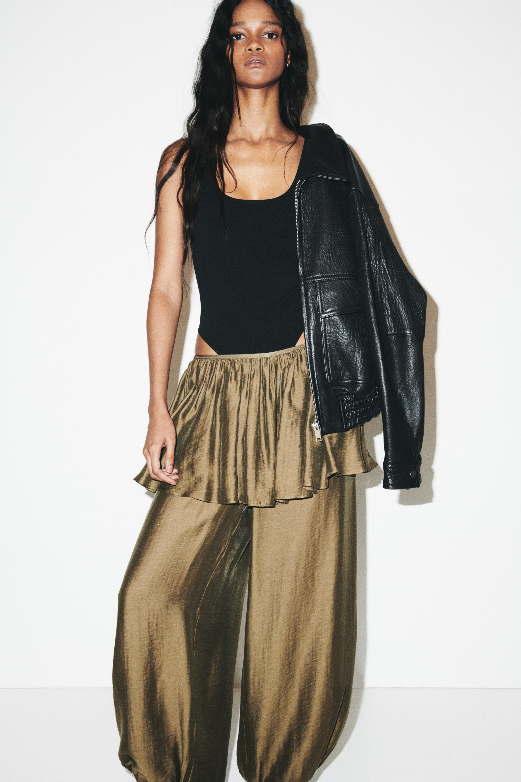 View larger image: The model is wearing a black sleeveless thong bodysuit with a scoop neckline, paired with metallic bronze wide-leg trousers and a black leather jacket draped over her left shoulder.