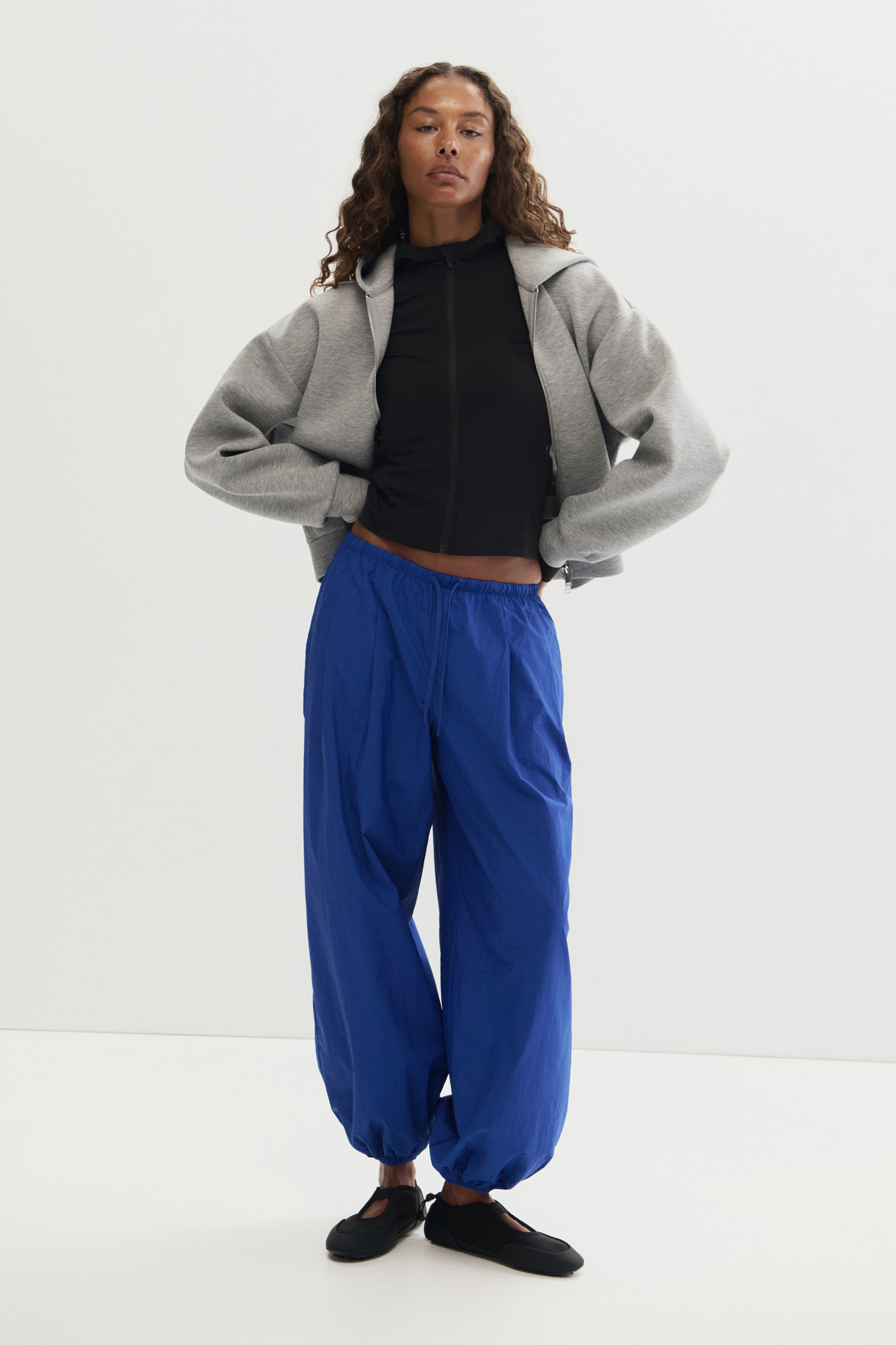 View larger image: Blue loose-fit activewear pants in water-repellent nylon fabric are worn by the model. The pants feature an elasticated drawstring waist, balloon-style legs, and elasticated hems, and are paired with a black zip-up top and a grey hooded jacket.