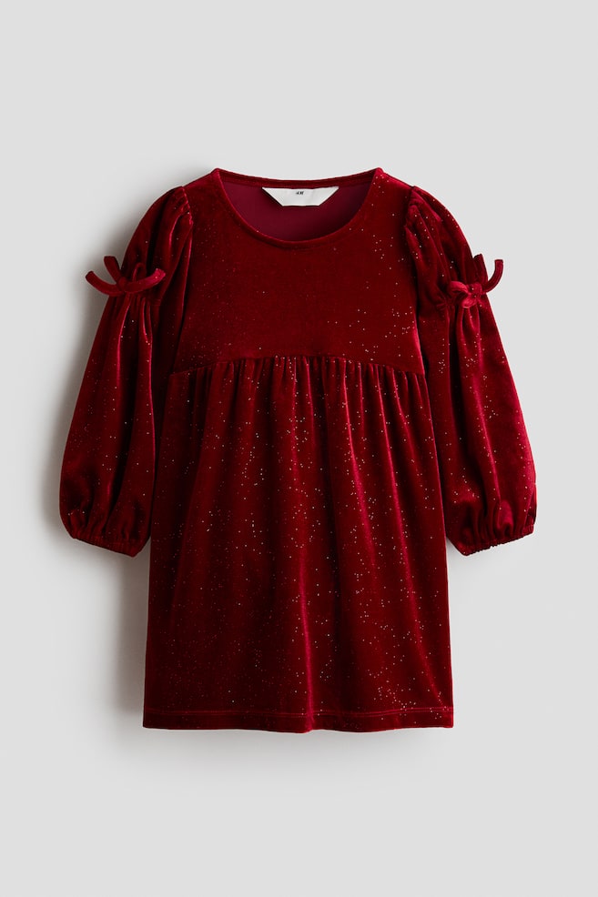 A deep red velvet dress is shown from the front, featuring a round neckline, an empire waist with a gathered skirt, and long puff sleeves with decorative bows on the upper arms. The soft velvet fabric is displaying scattered glitter throughout. - 1