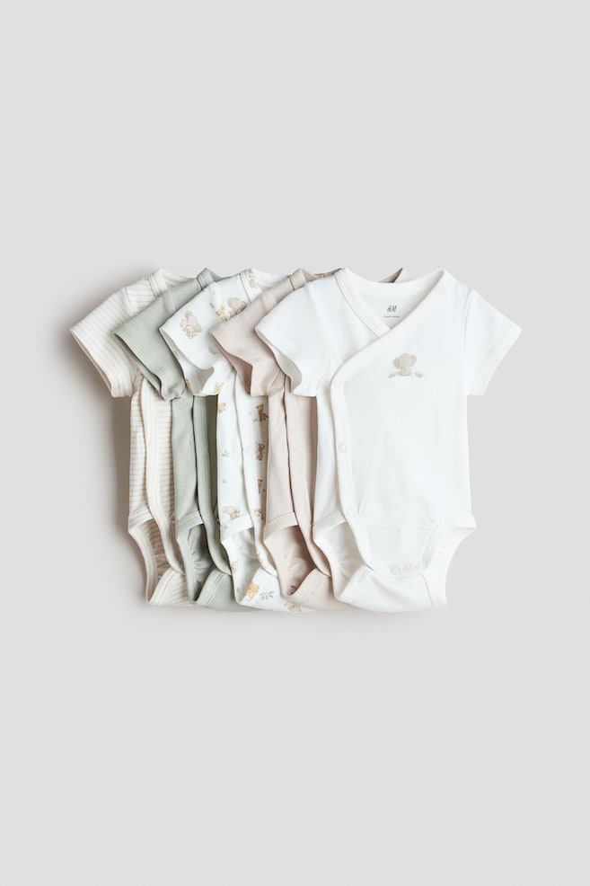 A stack of five short-sleeved bodysuits in soft cotton jersey is displayed with its front showing, featuring a wrapover design with snap closures at the sides and crotch. The bodysuits come in white, light green, and beige, with some showcasing subtle patterns like stripes or animal motifs. - 1