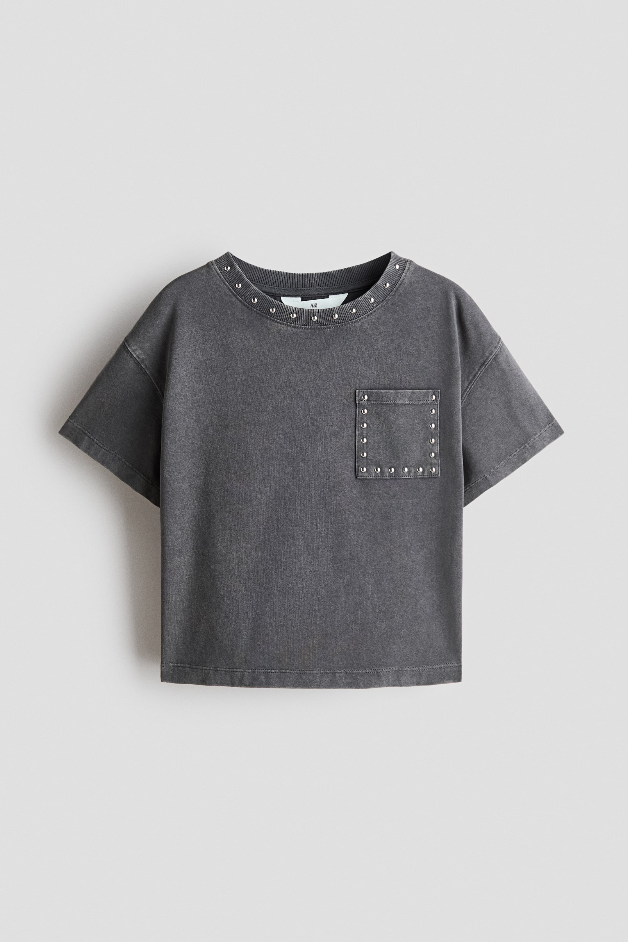 Ver imagen más grande: Una camiseta gris oscuro deslavada presenta su vista frontal, con un cuello redondo acanalado y mangas cortas. Pequeñas tachuelas plateadas decoran el escote y delinean un bolsillo de parche en el pecho izquierdo.