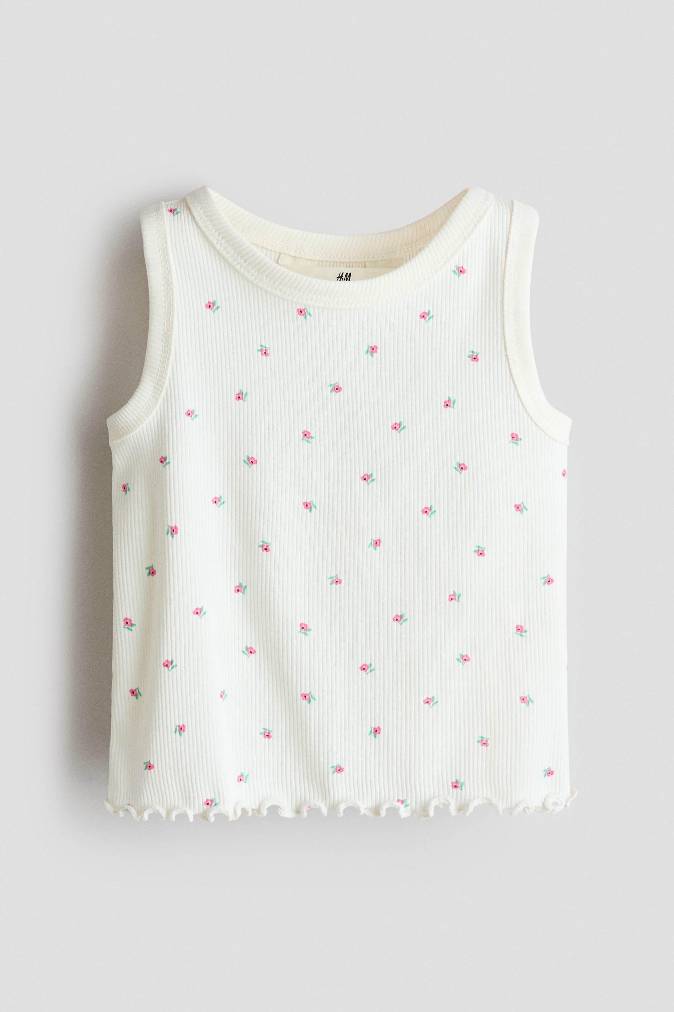 View larger image: A cream ribbed tank top is showcasing its front, adorned with a pattern of petite pink flowers with green stems. It features a round neckline, sleeveless design, and a wavy, lettuce-edge hem.