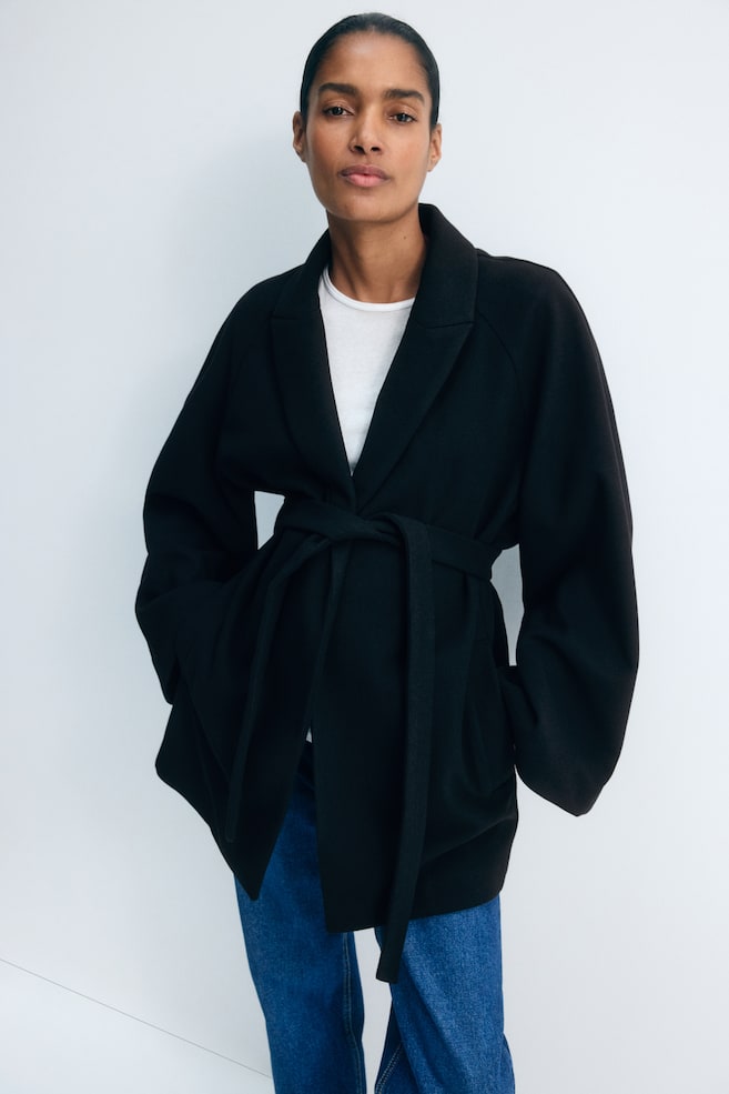 Um casaco preto em feltro, com lapelas em bico, mangas raglan compridas e cinto de atar a condizer na cintura, estÃ¡ a ser usado pela modelo. O casaco Ã© usado por cima de uma T-shirt branca e de jeans azuis, com a modelo em pÃ© e as mÃ£os nos bolsos laterais. - 1