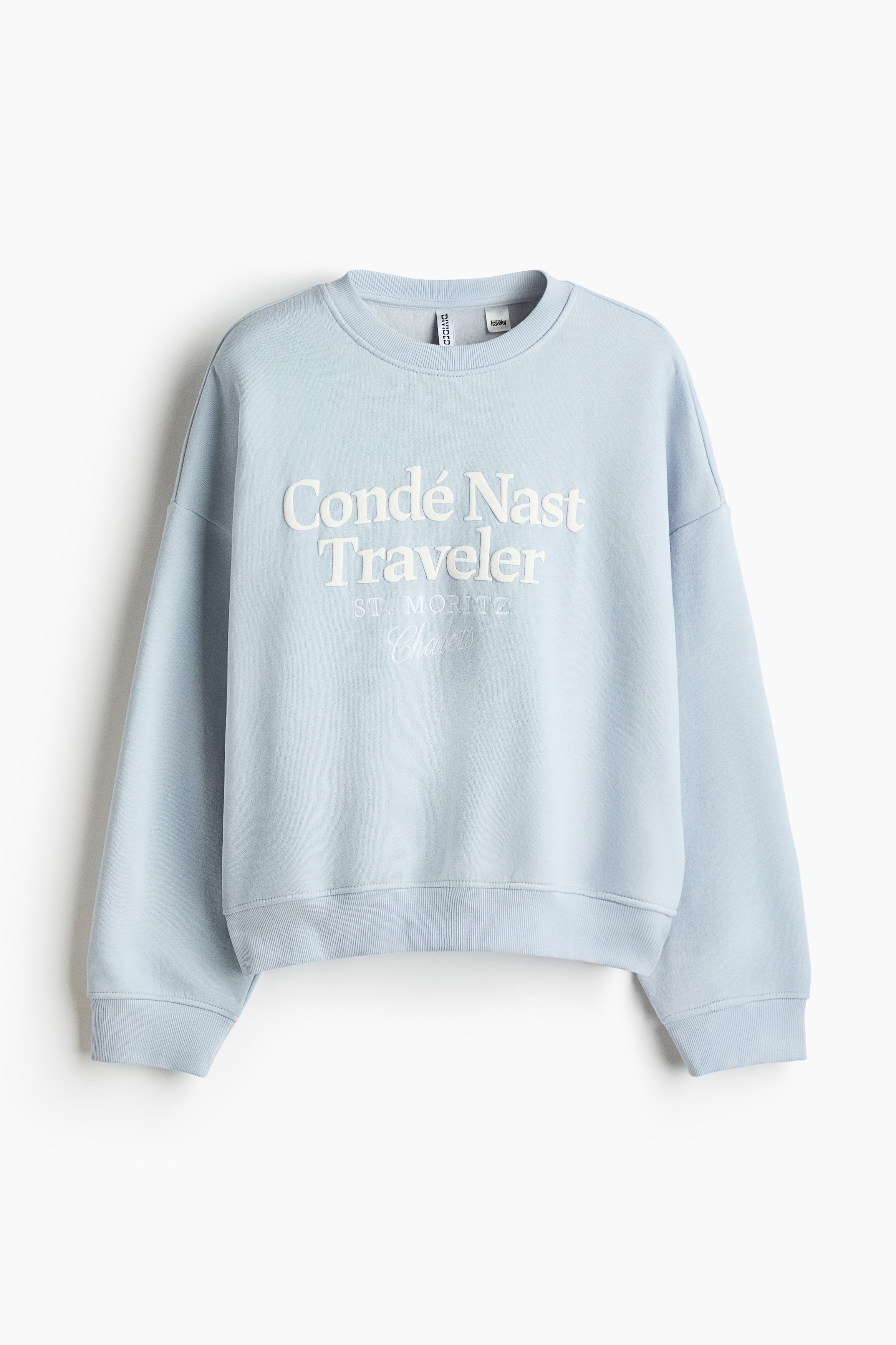 A light blue oversized sweatshirt is shown from the front, featuring a round ribbed neckline, dropped shoulders and long sleeves with ribbed cuffs. The sweatshirt features cream printed text reading âCondÃ© Nast Traveler ST. MORITZâ across the chest and is styled layered over a white long-sleeved top.