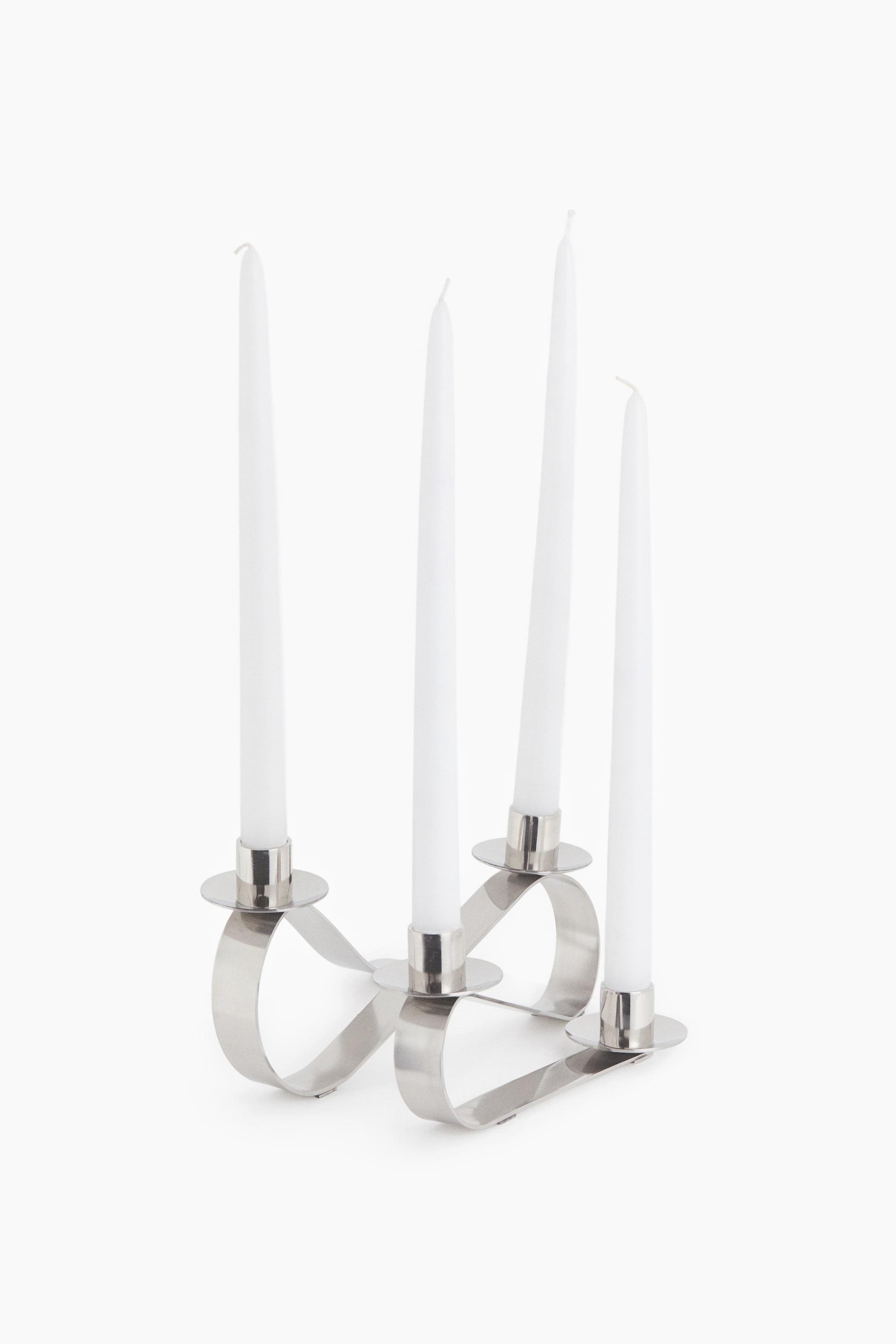 View larger image: A silver metal candelabra with a continuous, looping ribbon-like base holds four tall white taper candles. The product is shown front-facing, highlighting its sculptural, curved design, circular drip pans, and cylindrical candle cups.