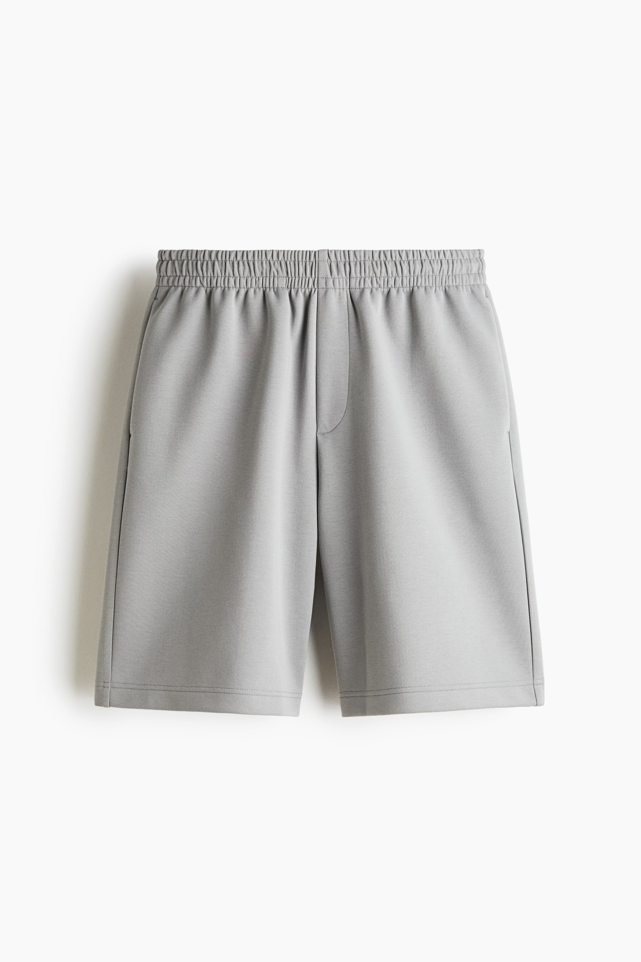 View larger image: Knee-length shorts in a light grey interlock sweat fabric are shown from the front, featuring a relaxed-fit silhouette. They display a smocked elastic waistband, a mock fly seam detail, and subtle side pockets.