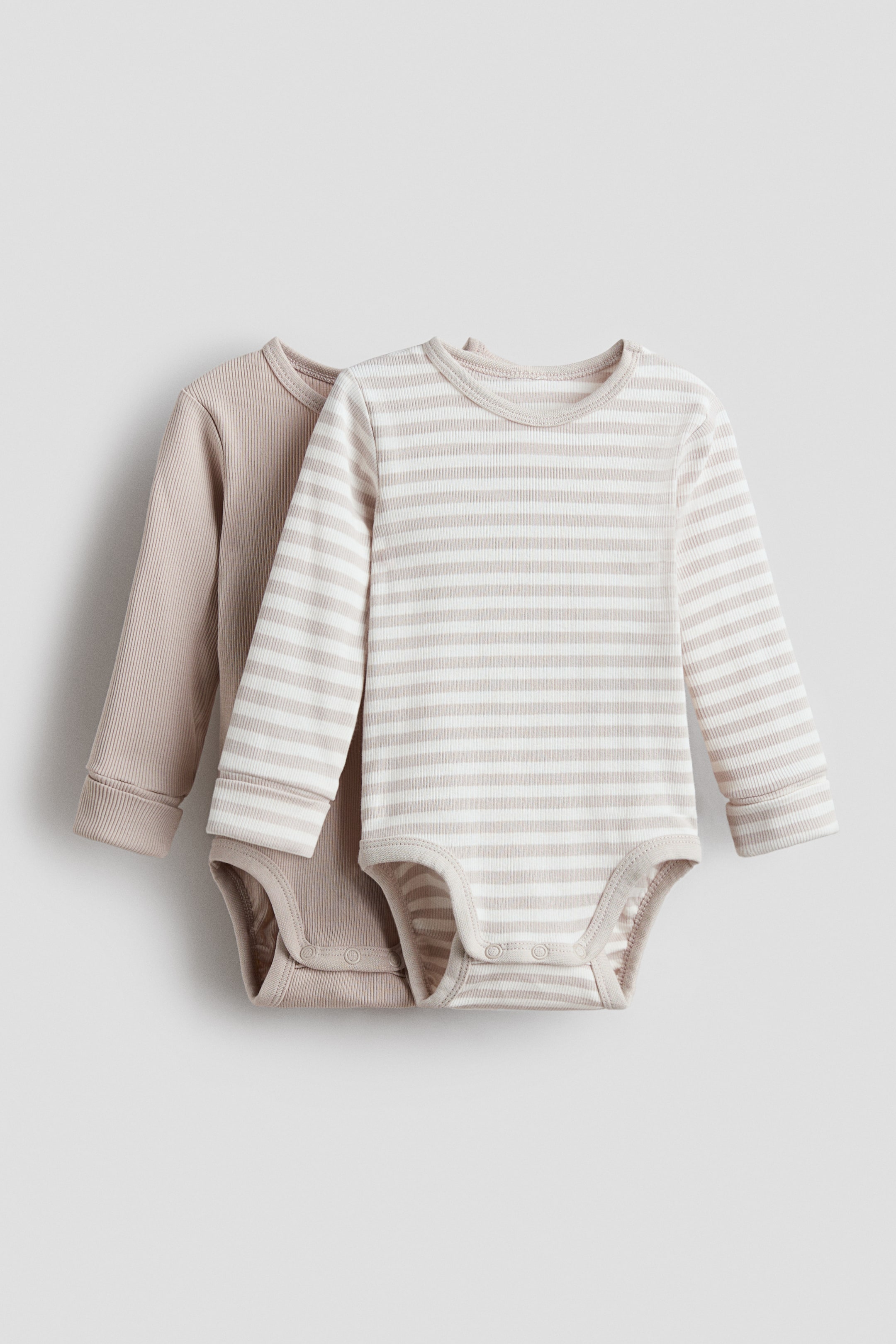 이미지 확대 보기: 긴 소매 아기 바디수트 두 벌이 평평하게 놓여 약간 겹쳐져 있으며, 골지 질감, 골지 커프스, 골지 크루넥을 보여줍니다. 앞쪽의 바디수트는 흰색과 연한 베이지색의 가로 줄무늬 패턴을 특징으로 하고 있으며, 그 뒤에 부분적으로 나타나는 것은 단색의 연한 베이지색입니다. 두 바디수트 모두 가랑이 부분에 스냅으로 고정되어 있습니다.