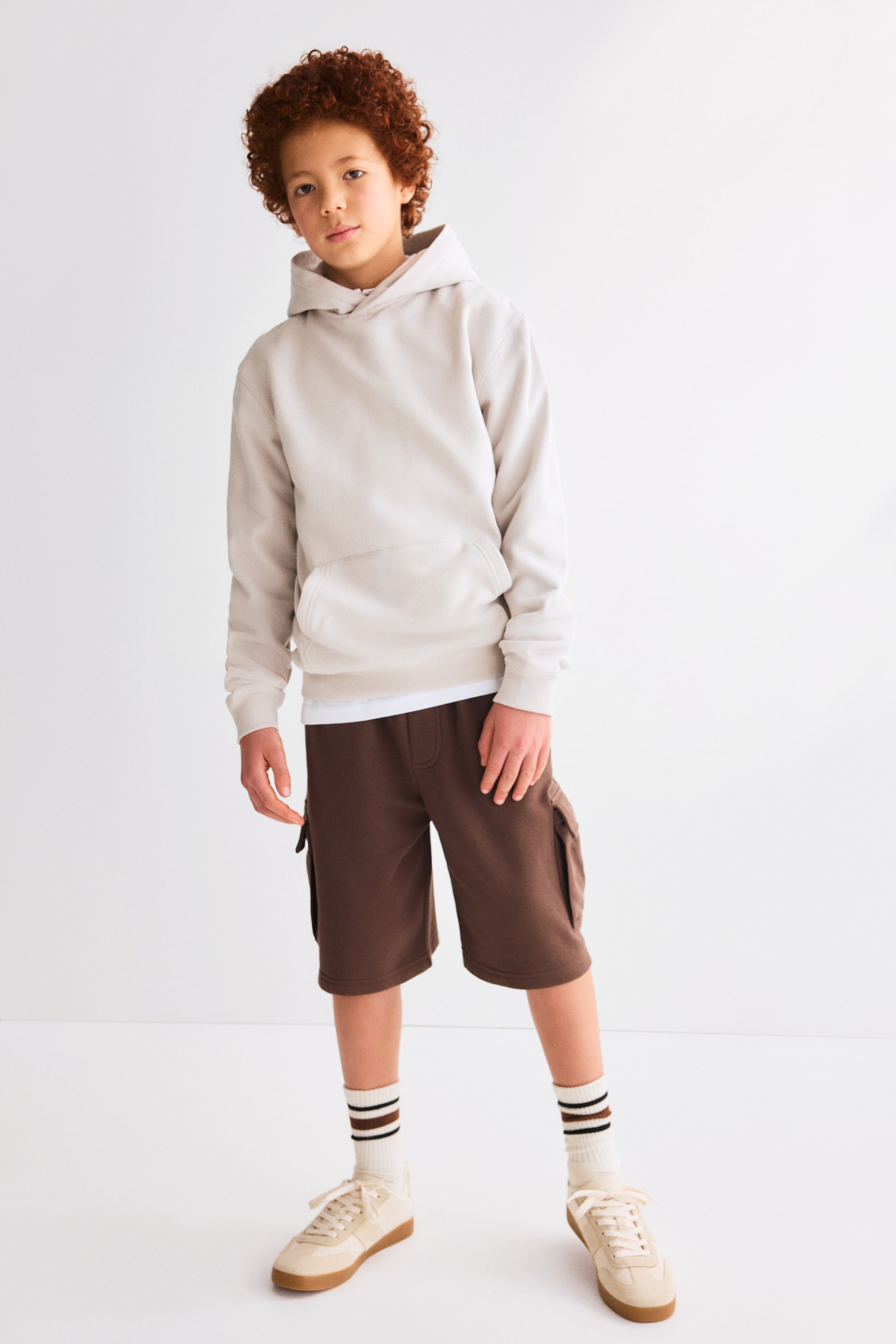 View larger image: A light beige hoodie is angled slightly to the right, showcasing a relaxed fit. It features a spacious kangaroo pocket at the front, complemented by ribbed cuffs on the sleeves and a ribbed hem at the bottom. A white t-shirt is layered beneath, peeking out from below the hem of the hoodie.