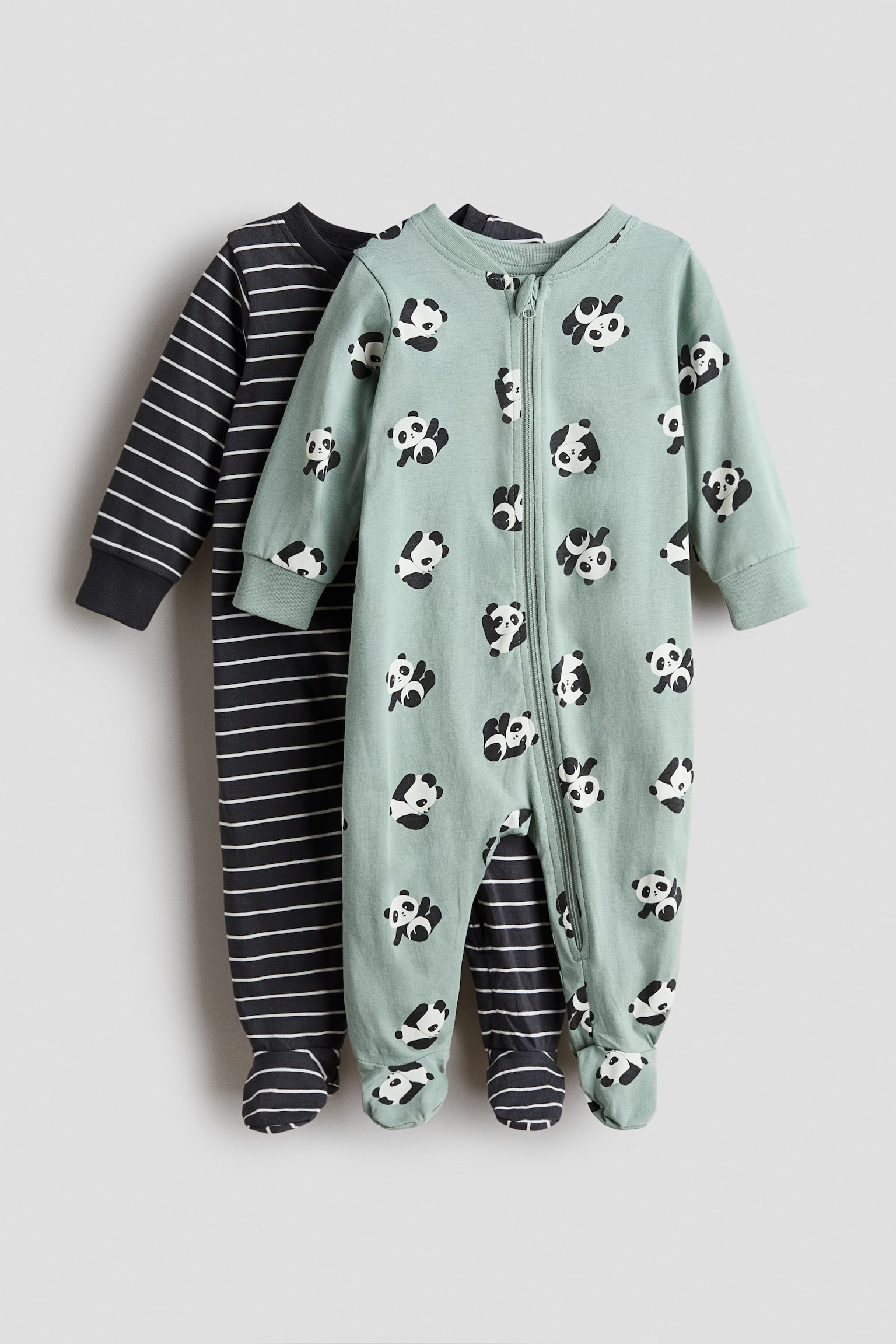 Twee babyrompers van katoenen jersey hangen in een aanzicht van voren, waarbij een lichtgroene romper met pandaprint over een donkergrijs gestreepte romper is gelaagd. Beide kledingstukken hebben lange mouwen met geribde boorden, gesloten voetjes en een ritssluiting aan de voorkant die doorloopt tot aan één pijp.