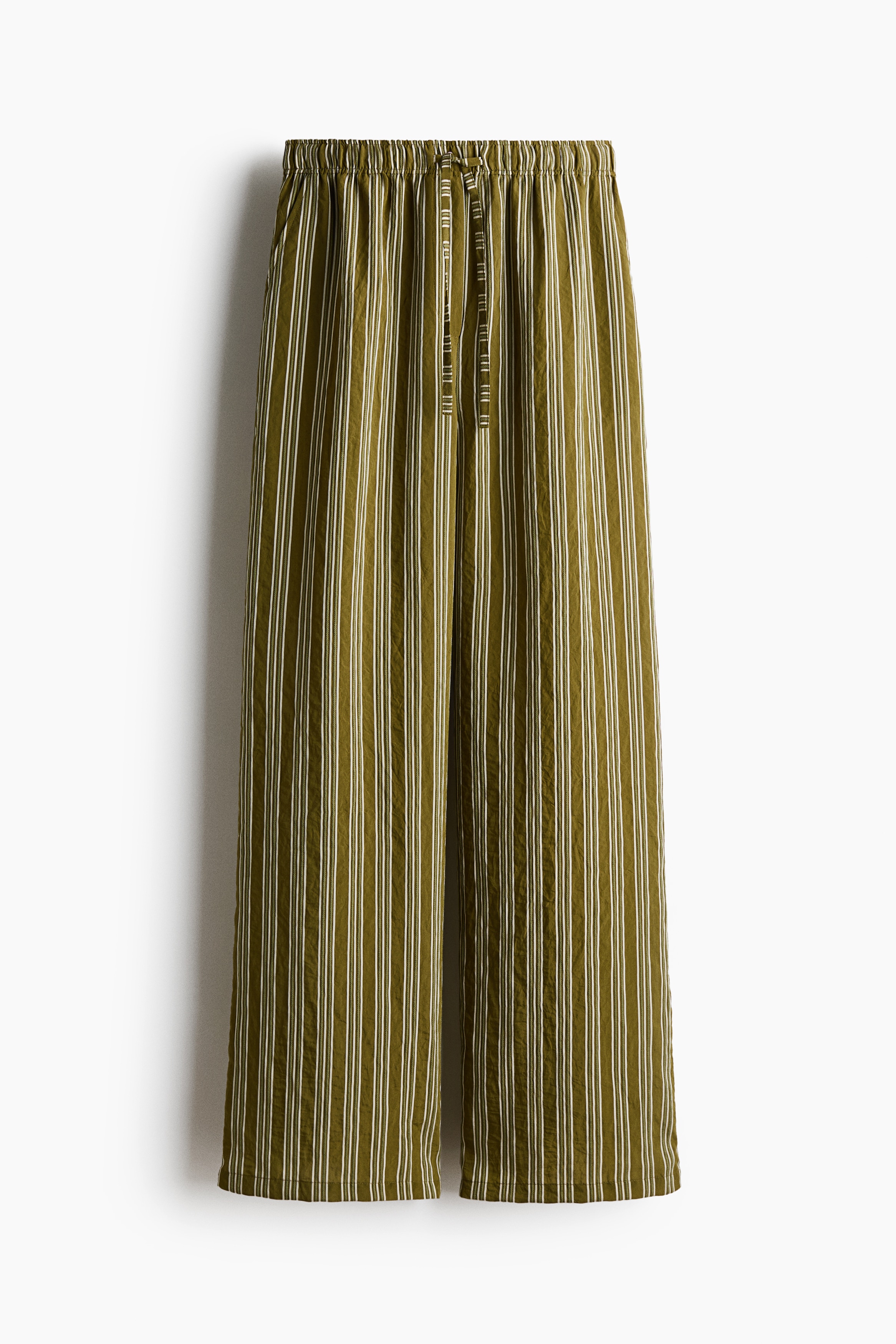Loose-fit, straight-leg pants featuring a vertical olive green and white striped pattern are worn by the model. The pants are styled with a matching long-sleeved striped tunic, belted at the waist, and black sandals.