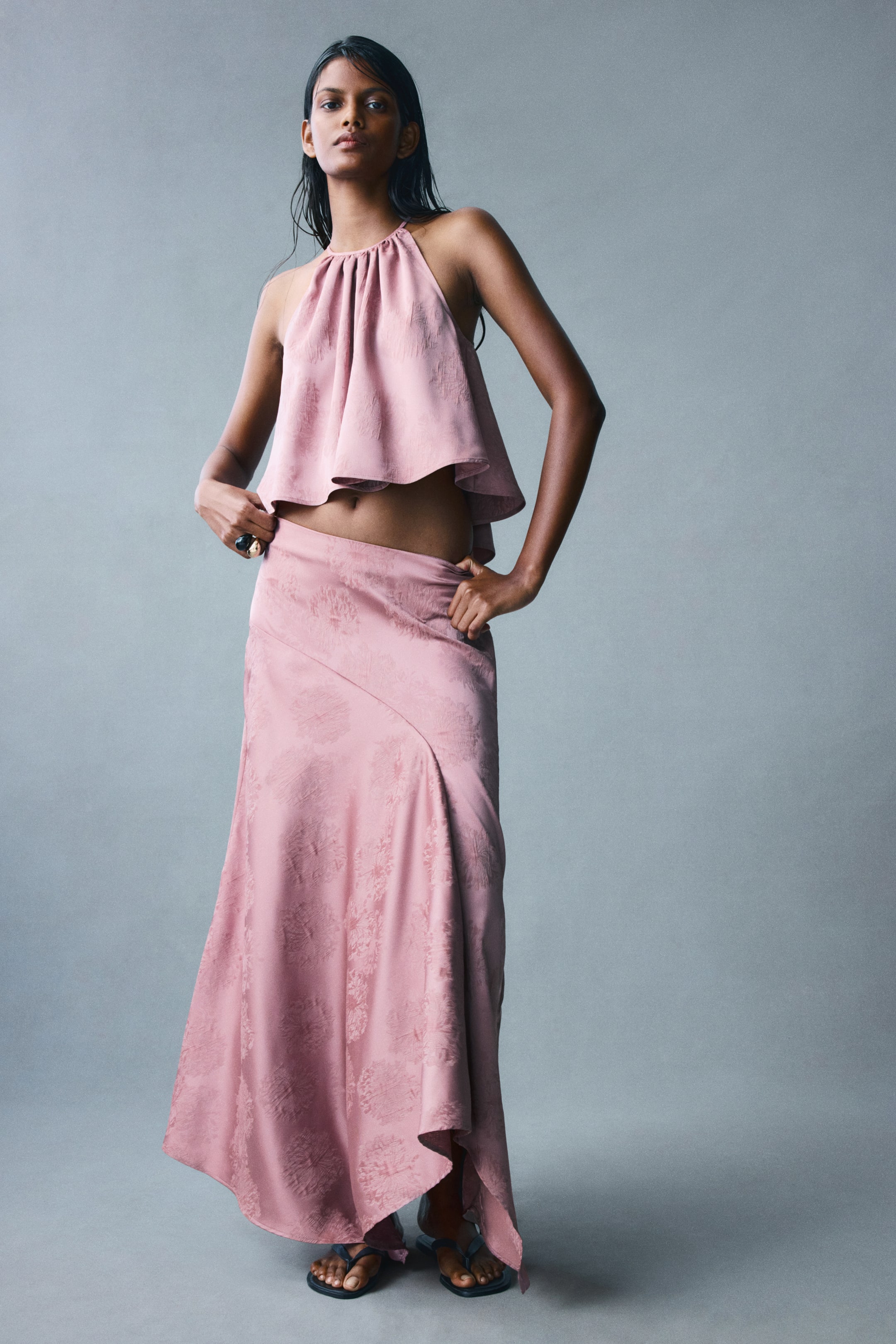 View larger image: The model is wearing a pink, calf-length satin jacquard skirt, featuring a diagonal seam across the front waist and a draped, asymmetrical handkerchief hem. The skirt is paired with a matching halter-neck top, with the model resting her hands near the waistband.