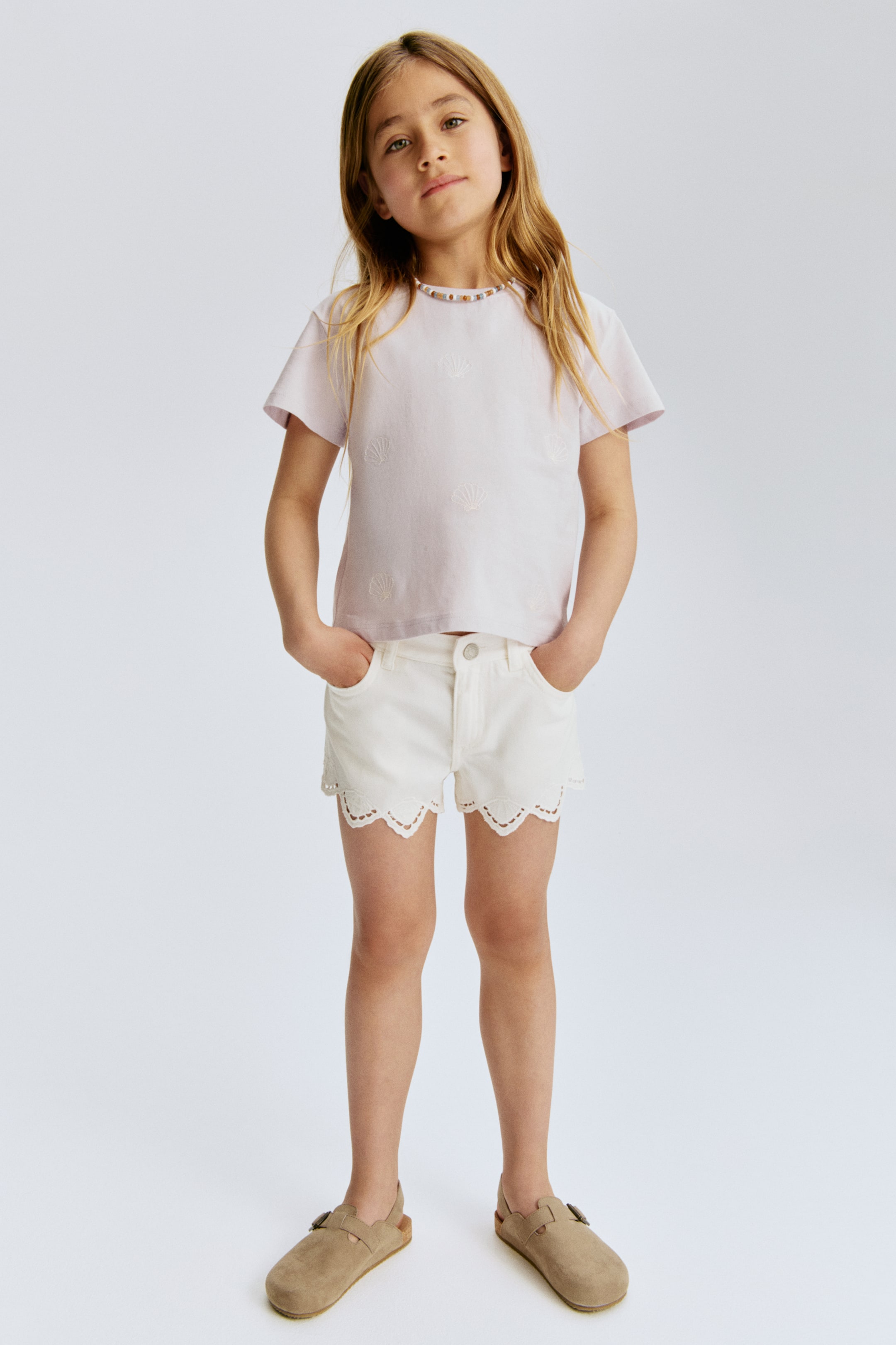 Größeres Bild ansehen: Ein helllila T-Shirt mit kurzen Ärmeln und gestickten Muschelmuster auf der Vorderseite, kombiniert mit weißen Shorts, die einen Muschelsaum mit Lochstickerei und Vordertaschen präsentieren. Hellbraune geschlossene Sandalen zeigen eine wildlederähnliche Textur, einen Schnallenriemen über dem Spann und eine korkähnliche Sohle.