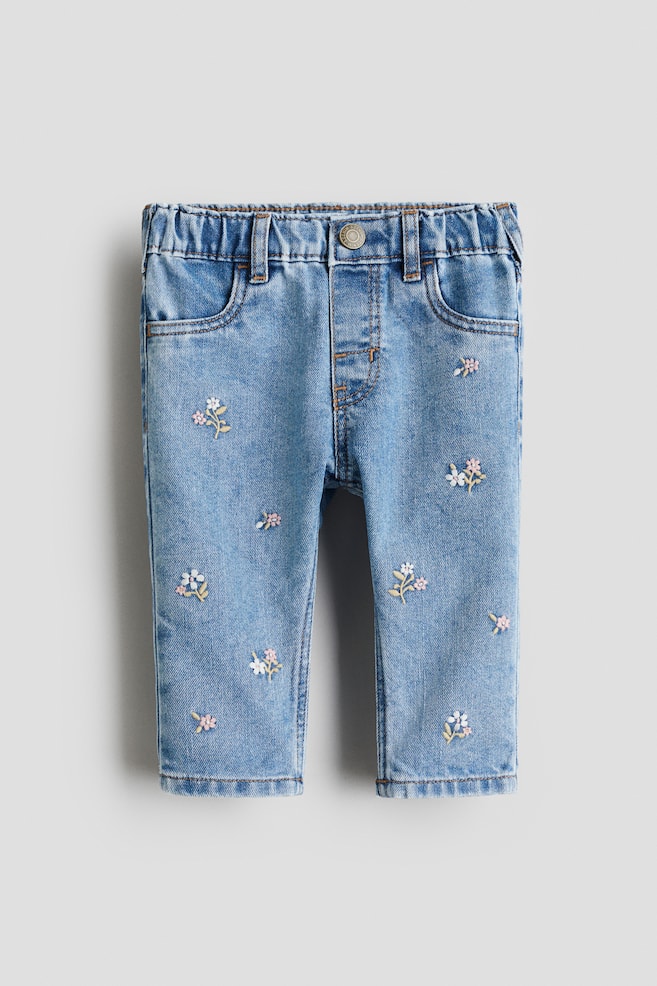 Light blue baby denim jeans are presented front-facing, featuring an elasticized waistband with belt loops, a single metal rivet button, and two front pockets. Scattered floral embroidery in pink, white, and yellow is adorning both legs. - 1