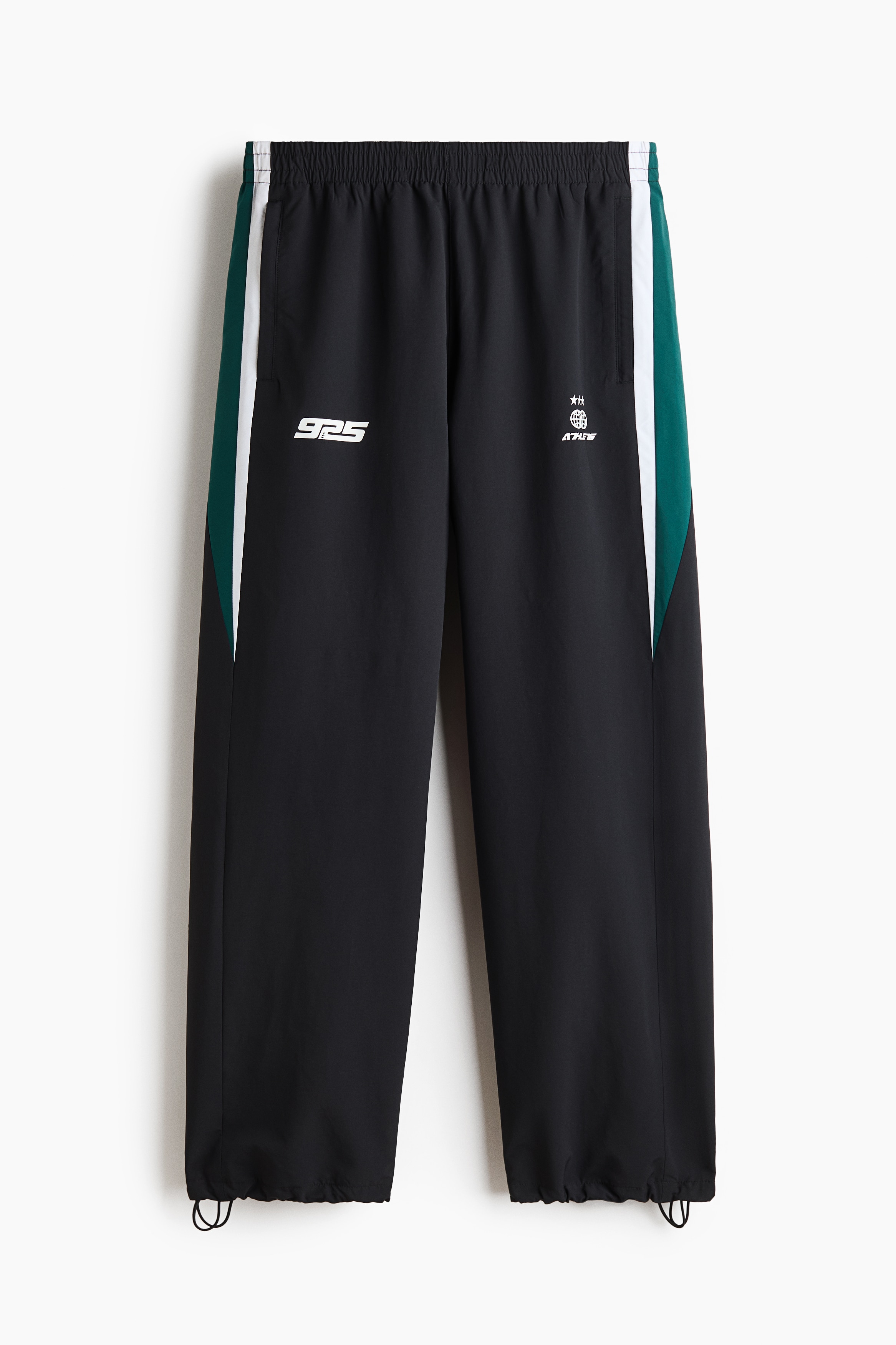 Relaxed-fit track pants in black woven fabric, featuring a white printed motif on the front thigh and a vertical teal and white side panel, are being worn by the model. The pants are styled with a coordinating color-blocked track jacket and light grey sneakers.