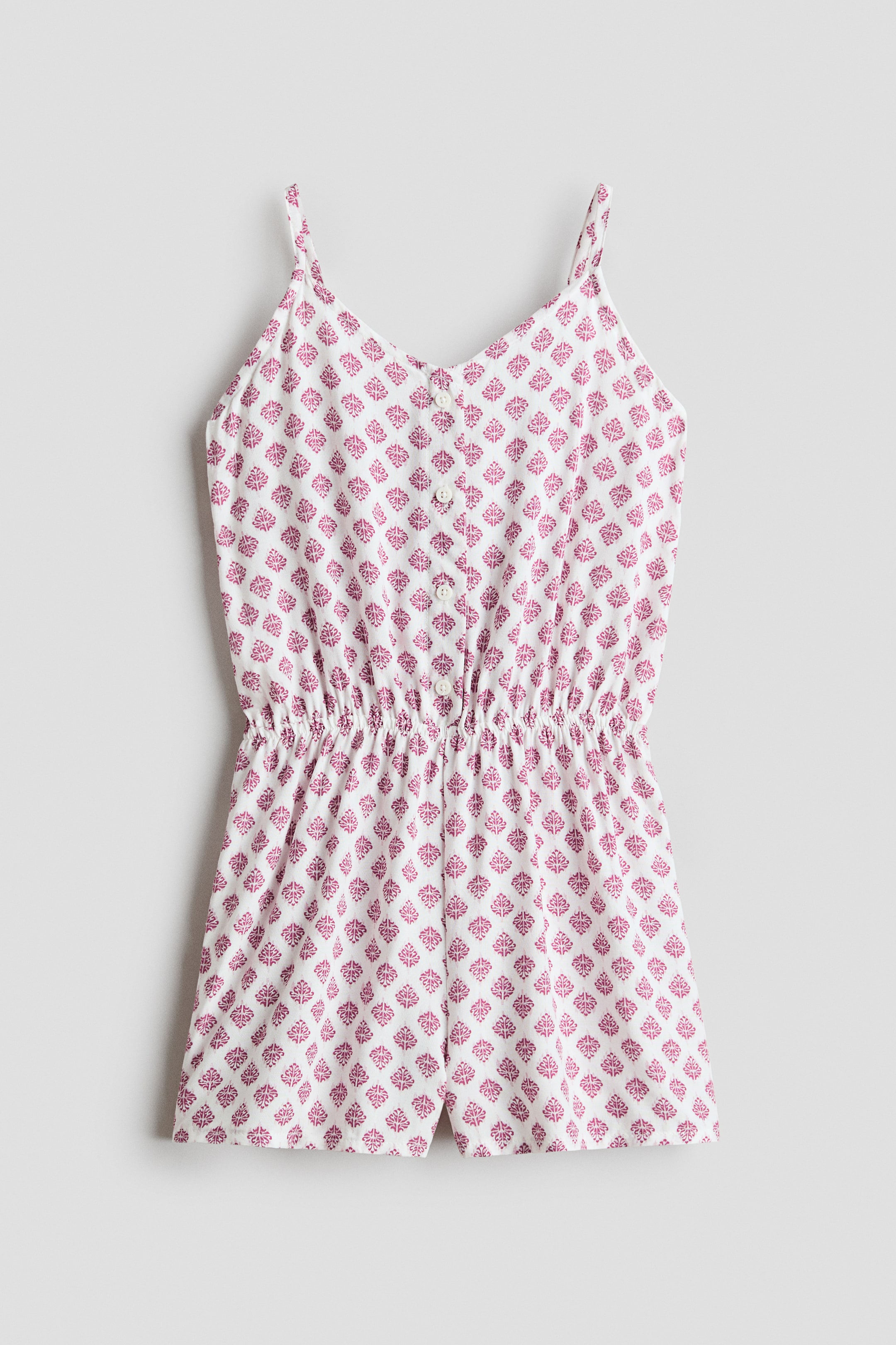 View larger image: A white romper, positioned front-facing, is showcasing a repeating magenta geometric pattern. It is designed with spaghetti straps, a V-neckline with a three-button placket, and an elasticized waist that is cinching the garment above shorts-style bottoms.
