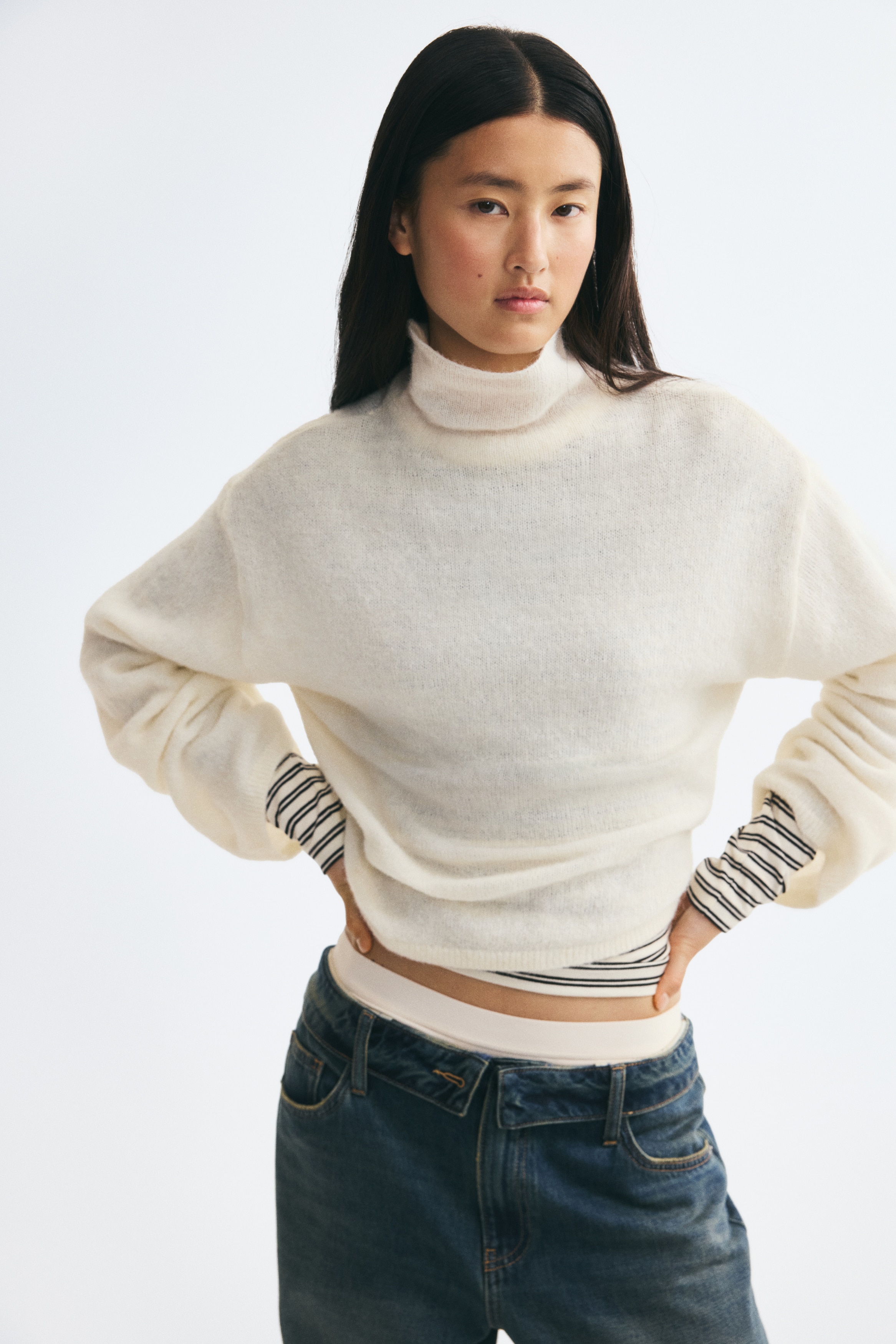 A cream-coloured knitted sweater is shown from the front, featuring a mock neck, dropped shoulders, and long, wide sleeves. The sweater showcases a soft, fuzzy knit texture and a relaxed, cropped silhouette.