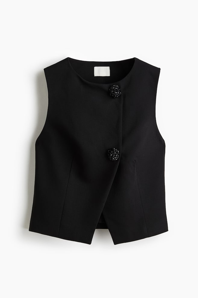 A black sleeveless vest is displaying its front, featuring a round neckline and an asymmetrical overlapping panel. Two large, round, black beaded buttons are adorning the vest's front. - 1