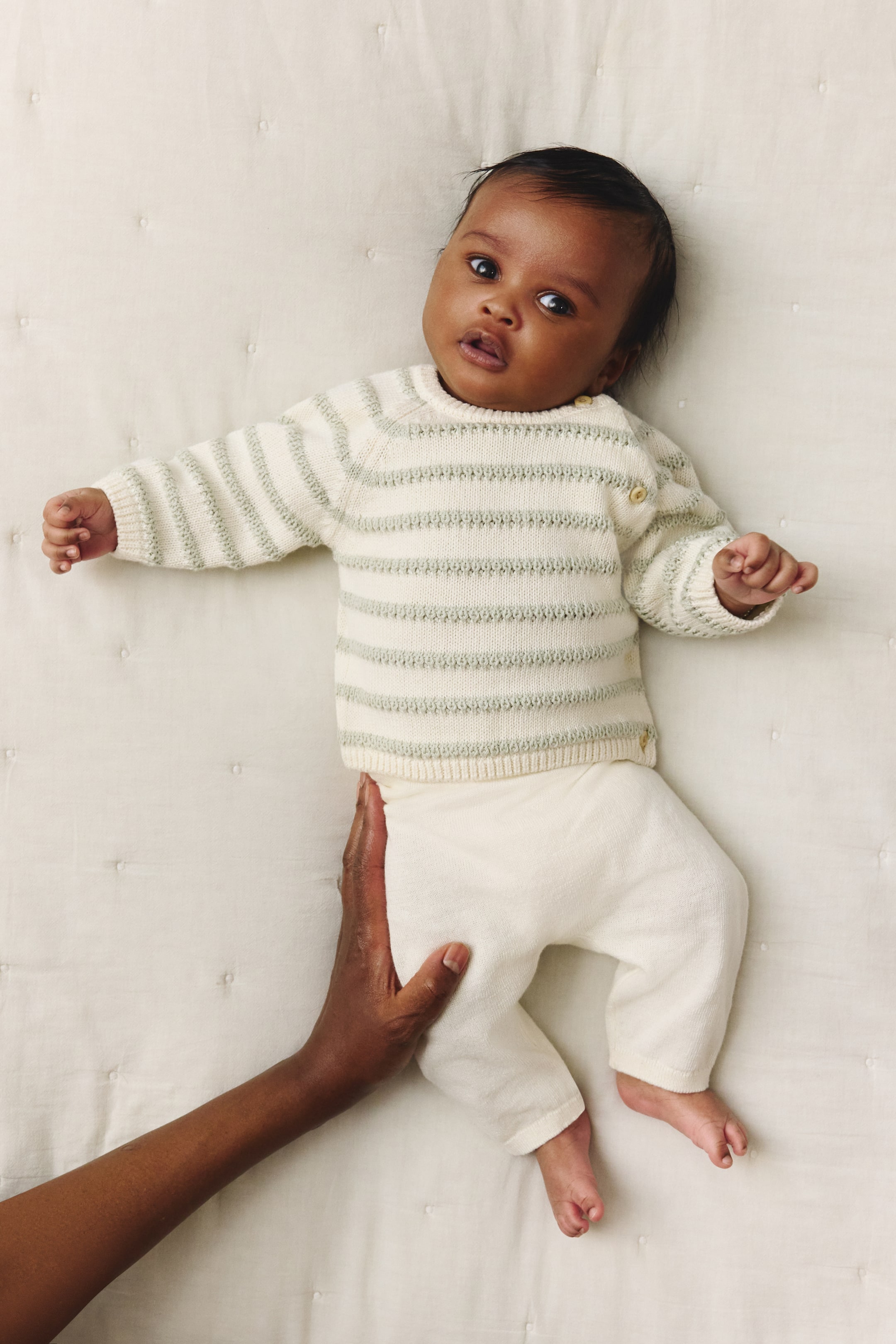 View larger image: A cream-coloured knitted jumper, featuring light green horizontal stripes, a round neck, ribbed cuffs, and small buttons on the left shoulder, is being worn. Matching cream-coloured knitted trousers are also being worn, with a hand gently holding the leg, showcasing their soft, relaxed fit.