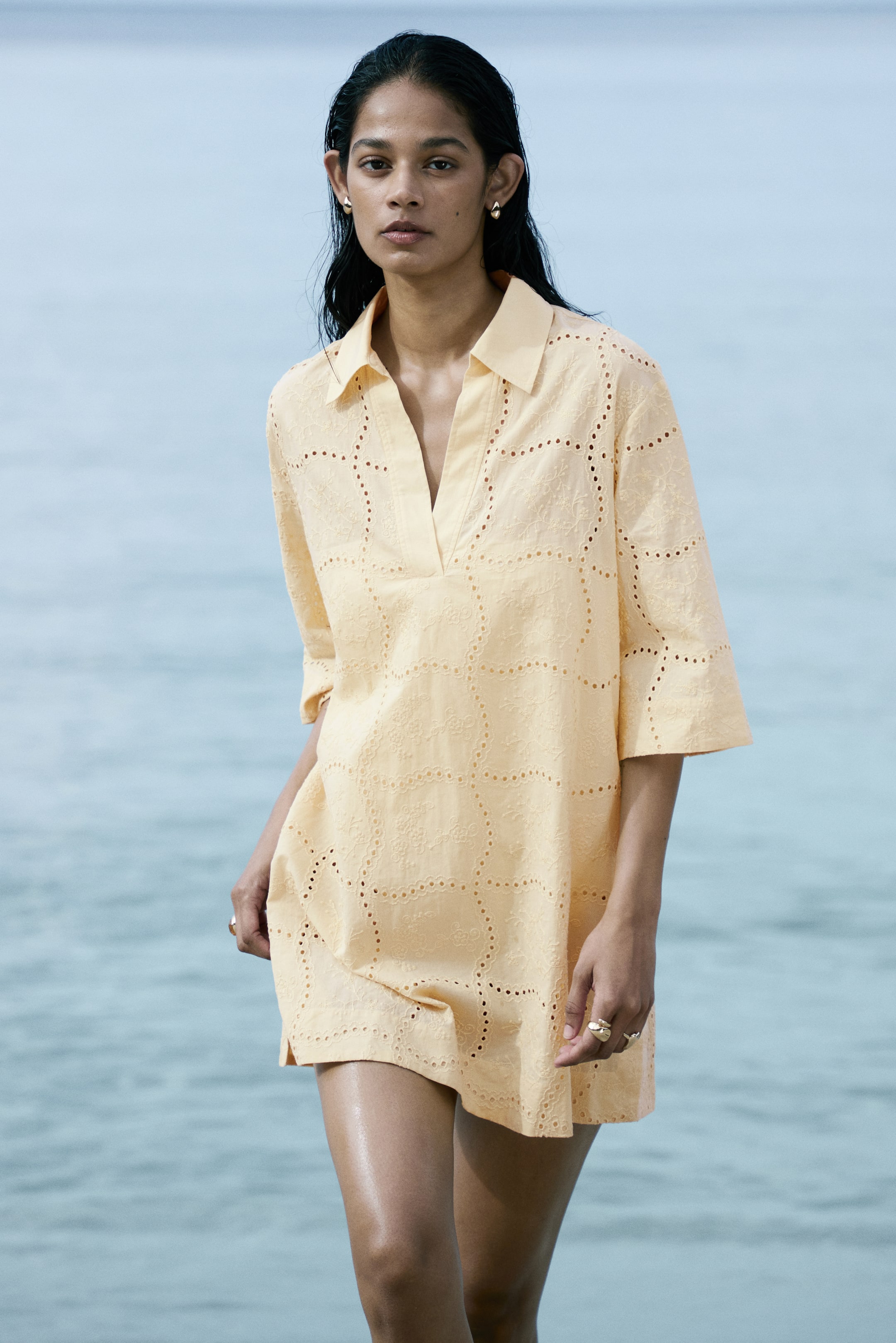 View larger image: A light yellow broderie anglaise tunic dress featuring a pointed collar, V-neckline, and three-quarter length sleeves, adorned with circular eyelet patterns throughout the fabric and small side slits at the hem.