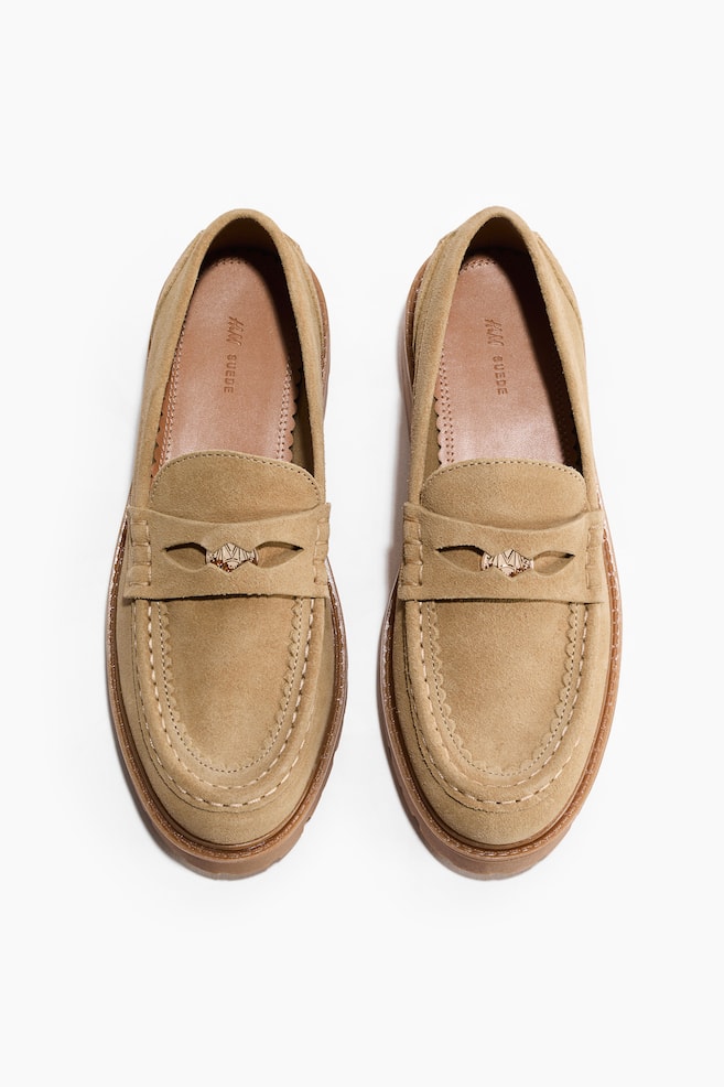 A pair of tan suede loafers is prominently displaying moc-toe stitching and a strap across the vamp, which is adorned with a small metallic ornament featuring an H&M logo. The visible insoles are showing "H&M SUEDE" text and scalloped edges. - 1