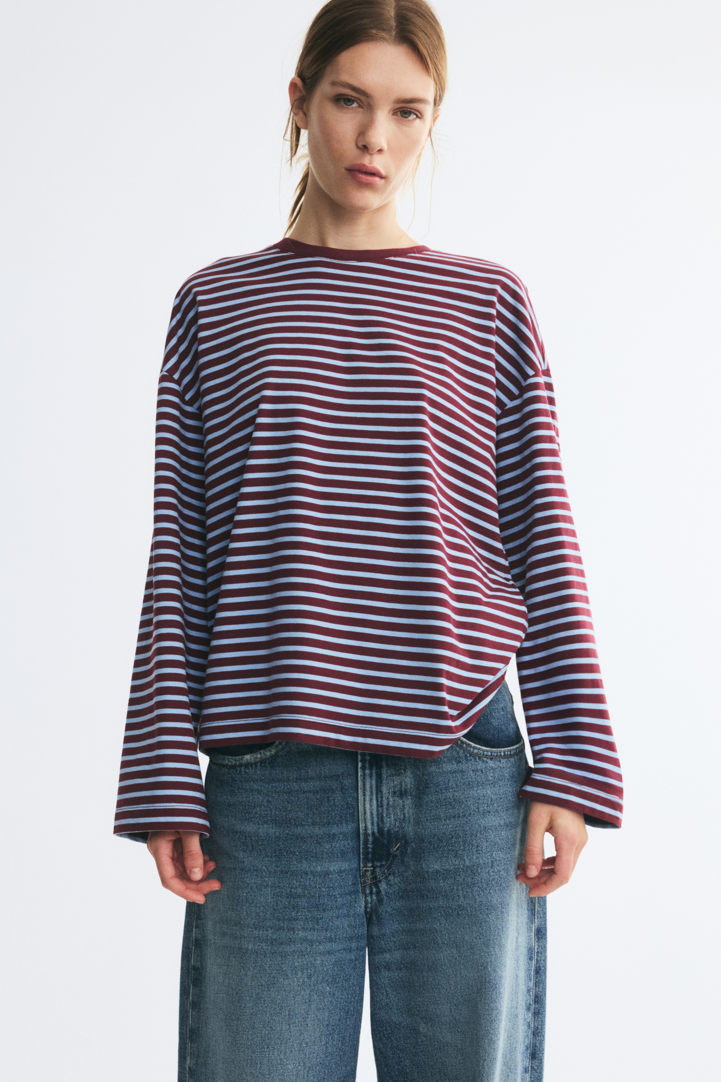Un top in jersey di cotone a maniche lunghe Ã¨ appeso in vista frontale, con righe orizzontali bordeaux e azzurro chiaro. Il capo presenta uno scollo rotondo tinta unita bordeaux, spalle scese e un orlo dritto.